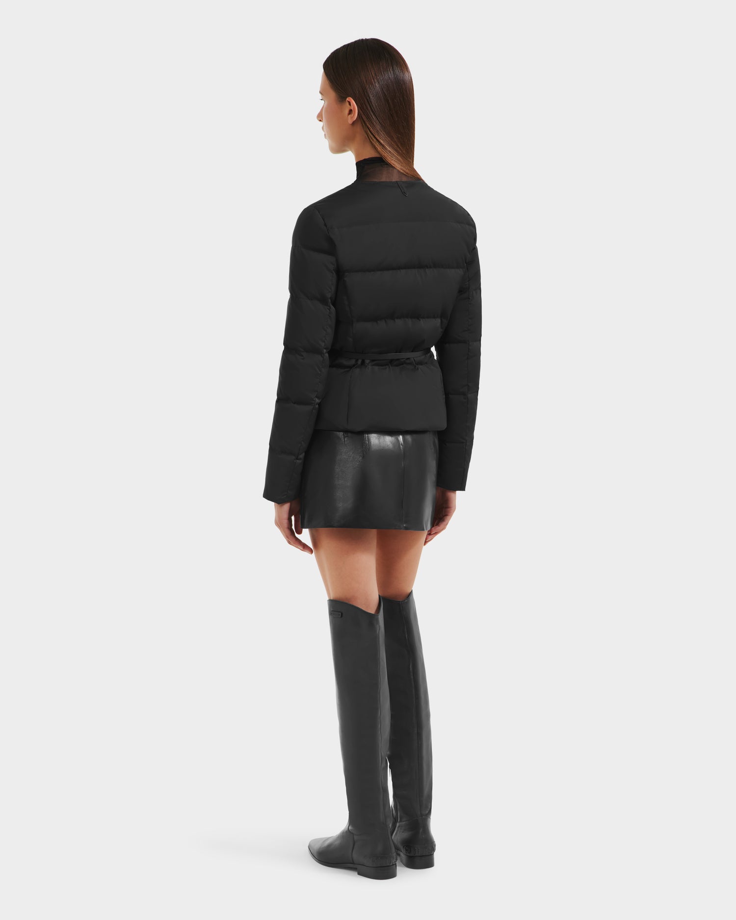 A woman faces away against a white background, wearing the Rudsak FRANCESCA - 8125021 Black puffer jacket with a short black leather skirt, vegan leather belt, and knee-high black boots.