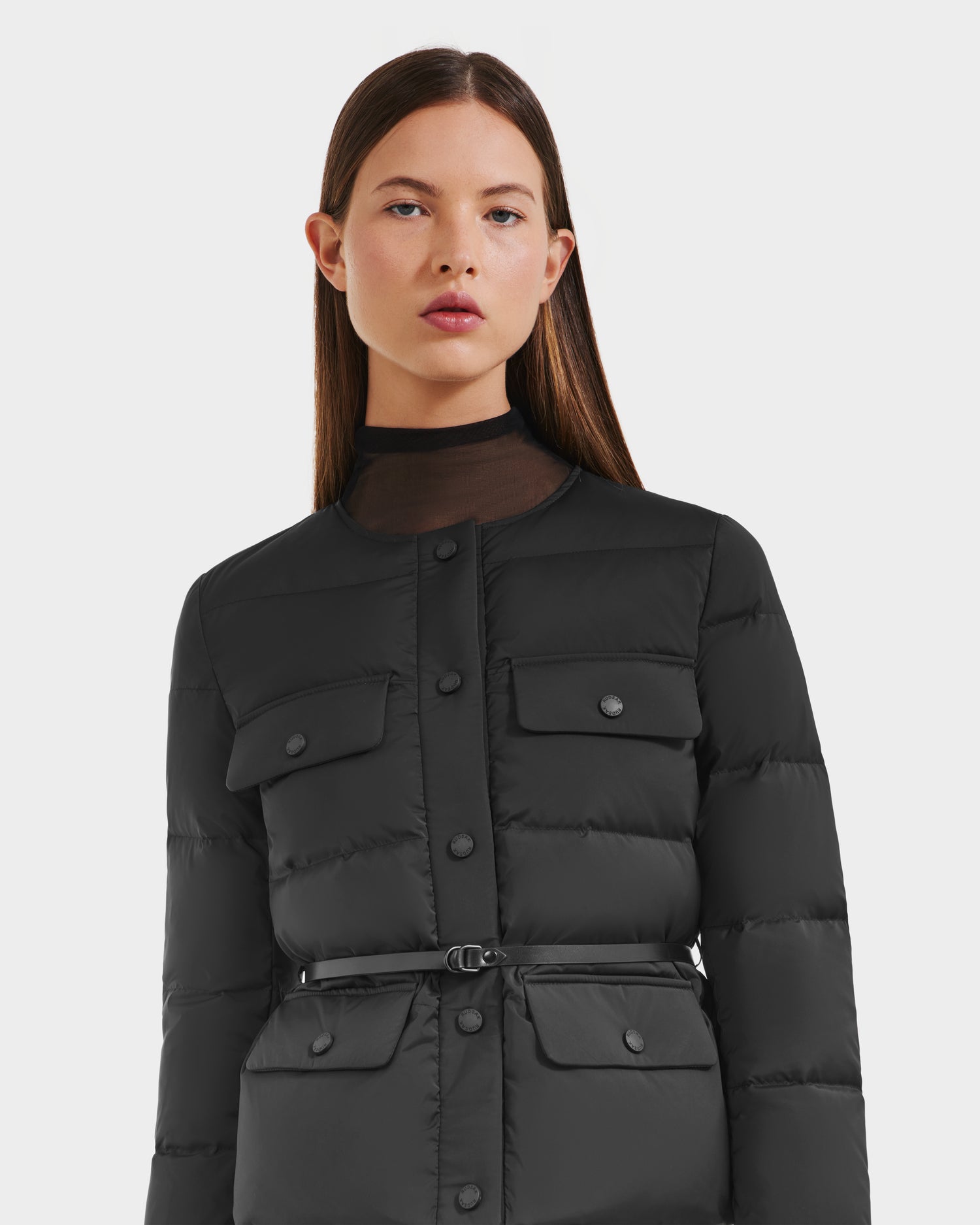 A woman with straight brown hair models the Rudsak FRANCESCA - 8125021 Black puffer jacket, featuring buttons, a vegan leather waist belt, and two chest pockets over a sheer black turtleneck against a light background.