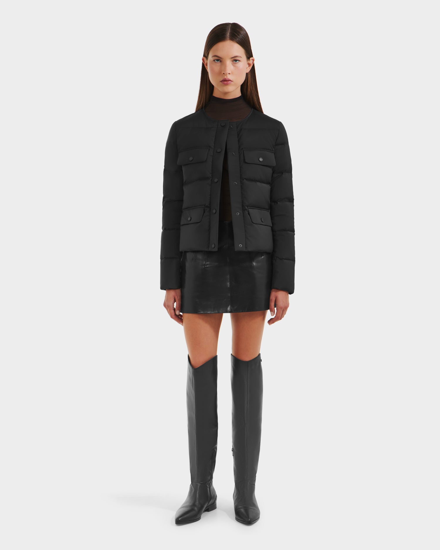 A woman faces forward against a white backdrop, wearing the Rudsak FRANCESCA - 8125021 Black lightweight puffer jacket with a black leather mini skirt, sheer black top, and black knee-high boots.