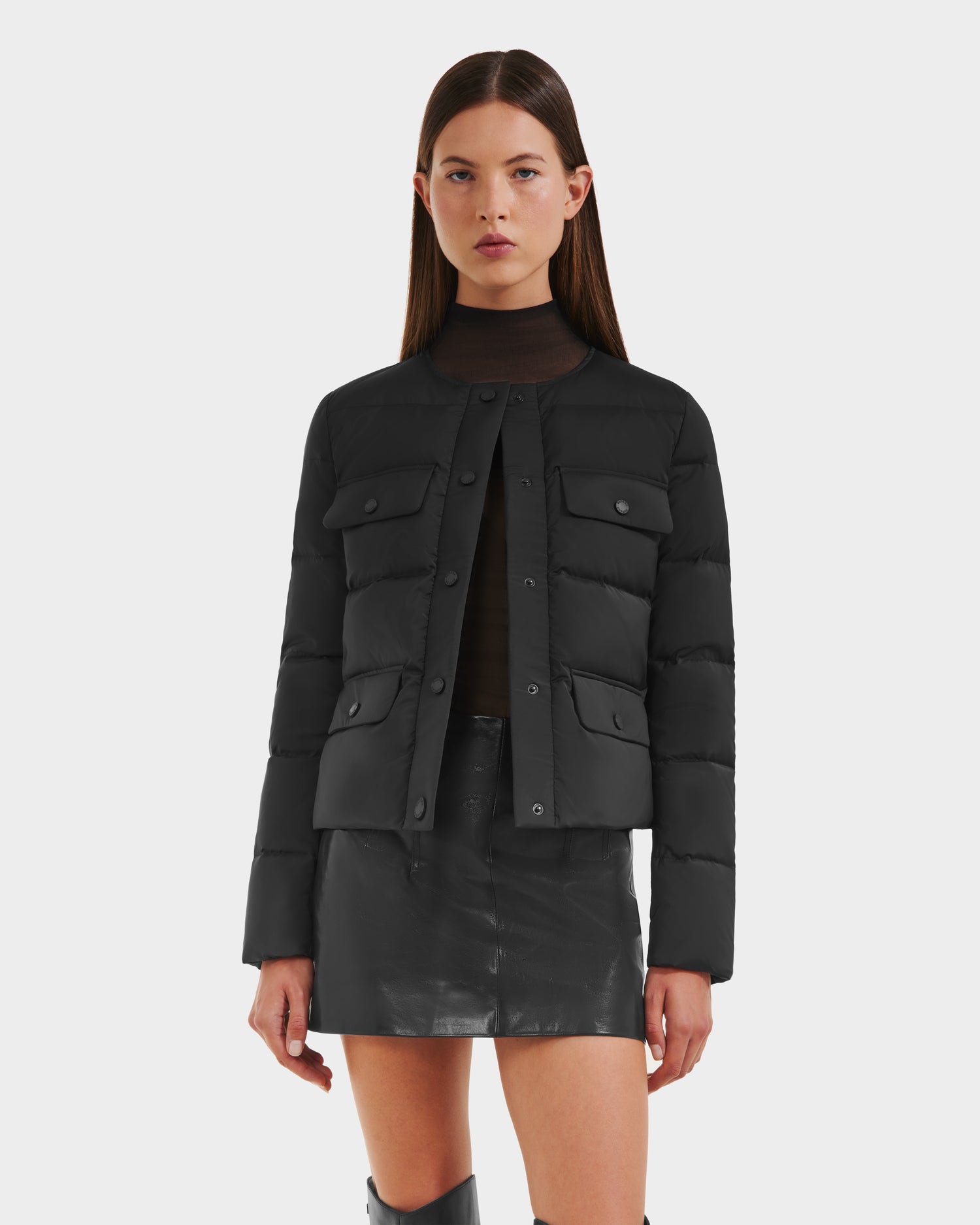 A woman with straight brown hair wears the Rudsak FRANCESCA - 8125021 Black collarless quilted puffer jacket over a dark top and black leather mini skirt, styled with a vegan leather belt, against a plain white background.