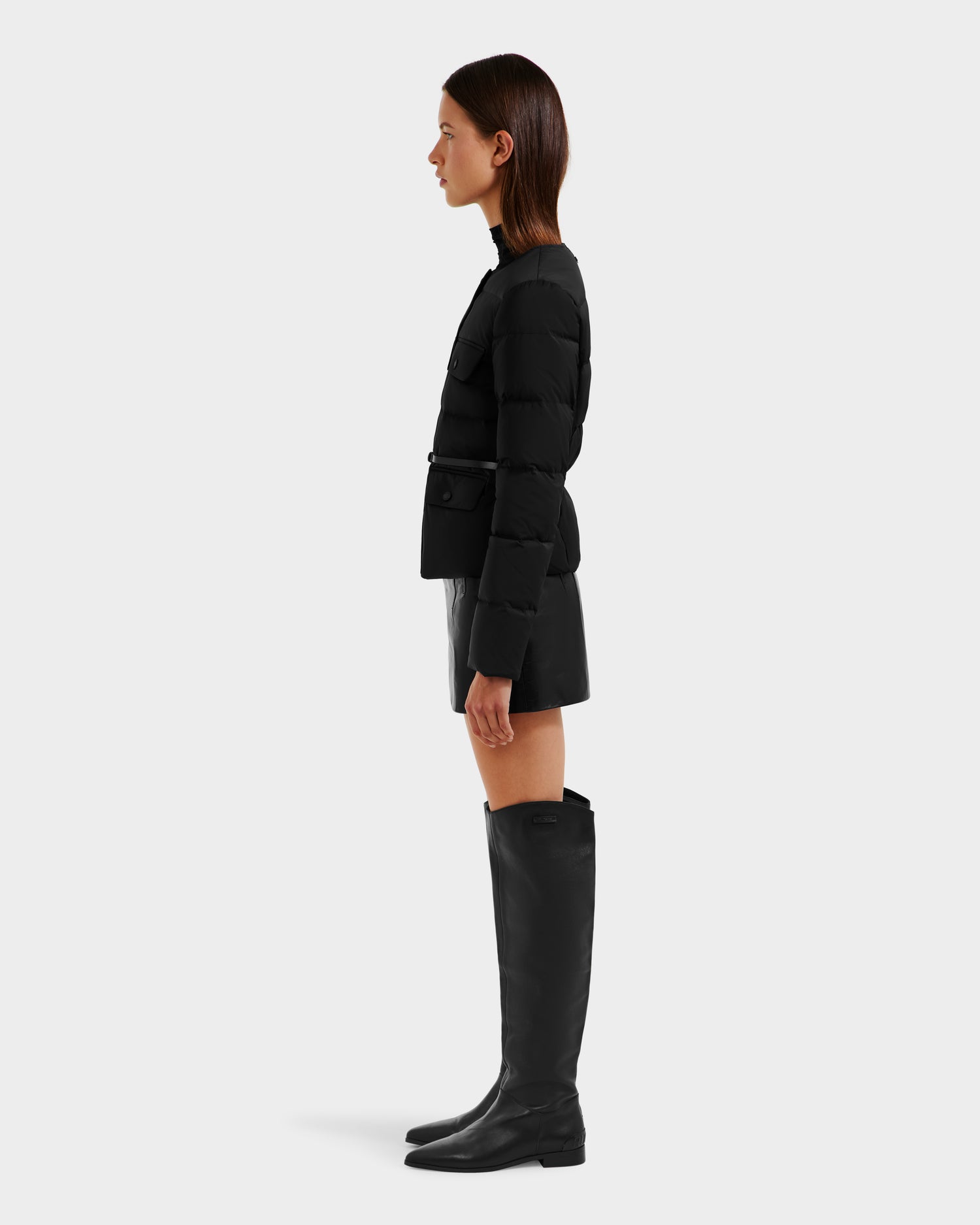A woman stands in profile against a plain background, wearing the Rudsak FRANCESCA - 8125021 Black quilted puffer jacket, paired with a black mini skirt and tall black boots.