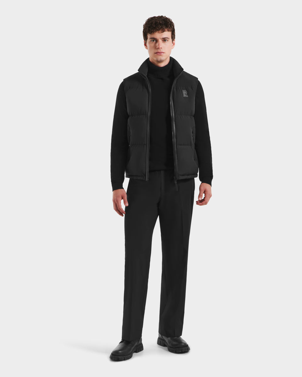 A man faces forward, modeling the Rudsak FREDDY - 6125571 Black down men’s puffer vest over a black long-sleeve shirt, trousers, and shoes, highlighting stylish lightweight layering against a plain white background.