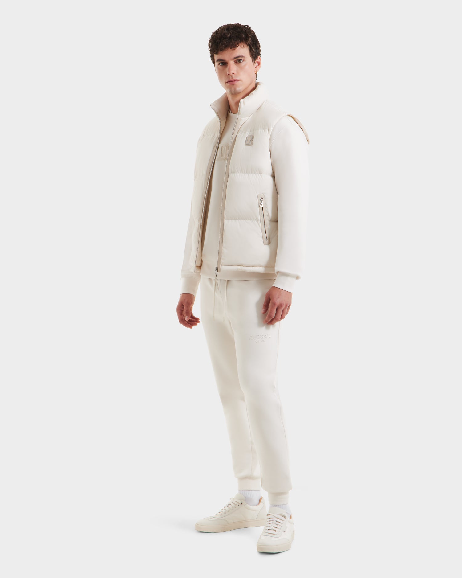 A man stands against a plain white background wearing the Rudsak FREDDY - 6125571 Vanilla mens puffer vest in cream over matching loungewear with white socks and sneakers, perfect for lightweight layering.