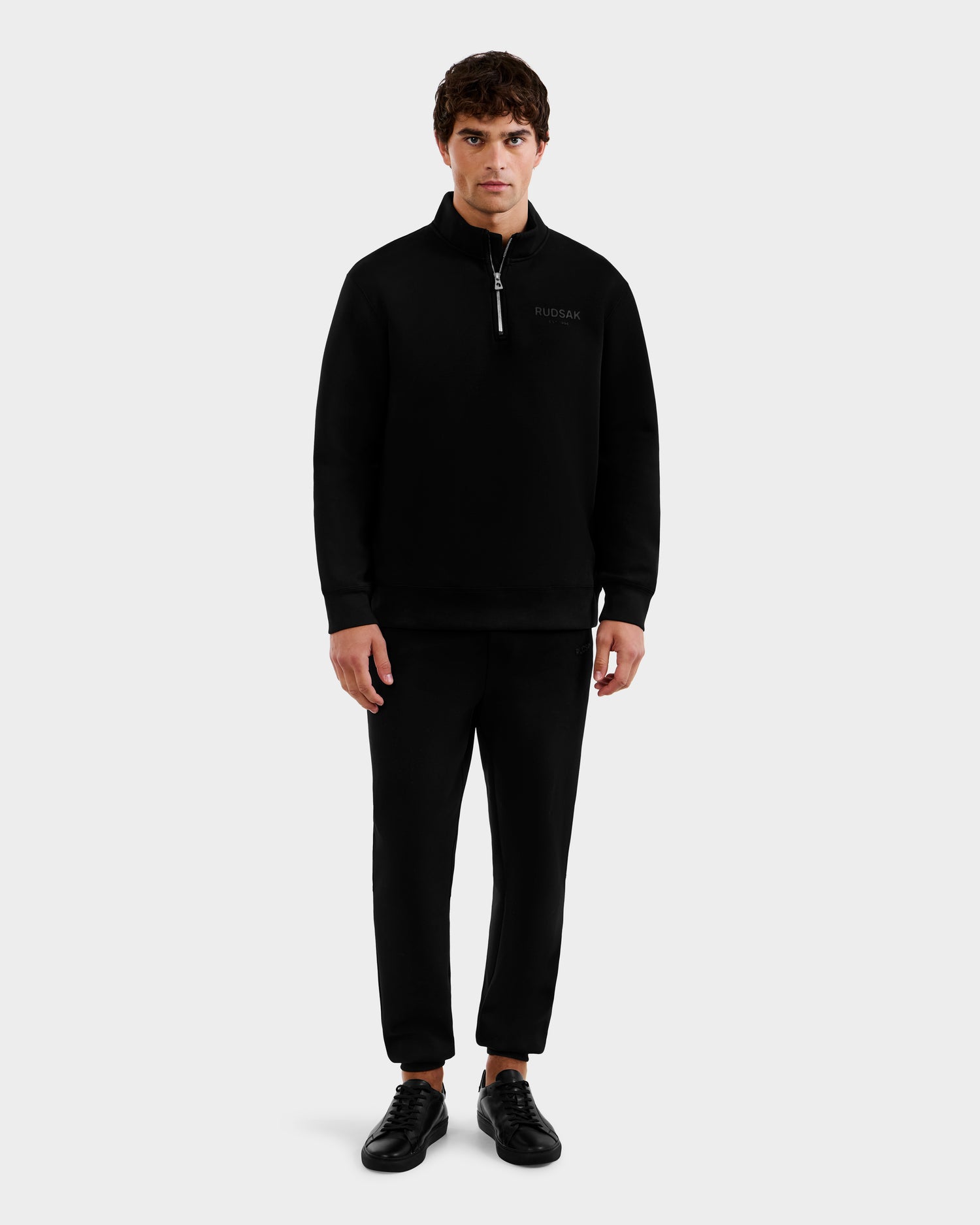 A man faces forward against a light gray background, wearing the Rudsak FREMONT - 9525000 Black M cotton mock neck sweater, paired with black pants and black sneakers.