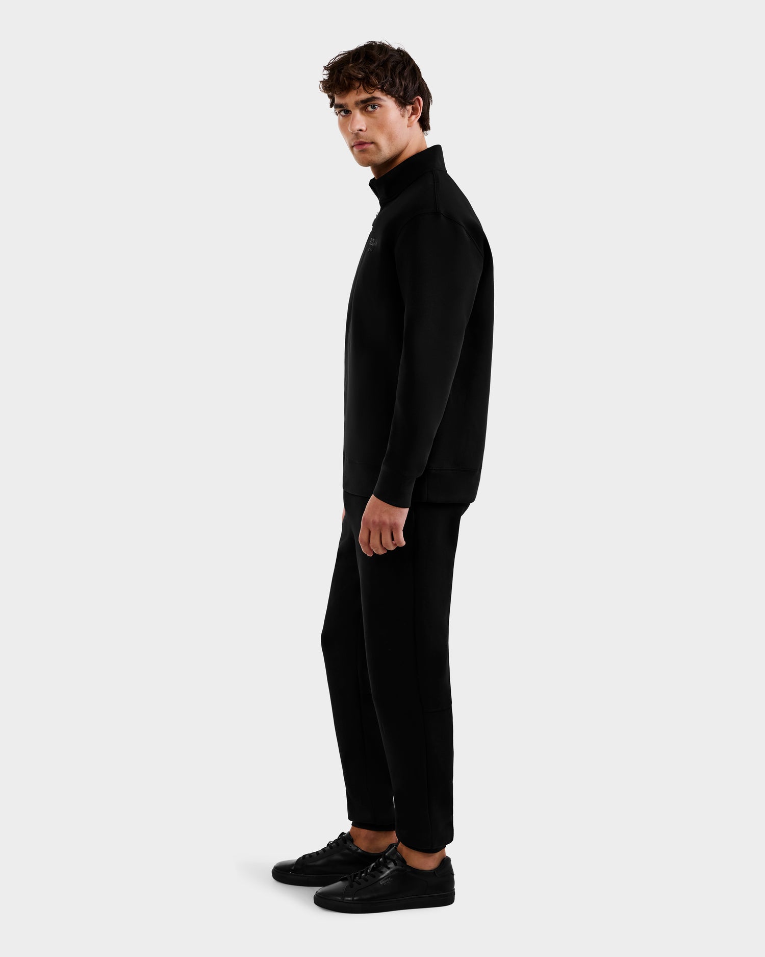 A man stands sideways against a plain light background, wearing the Rudsak FREMONT - 9525000 Black M sweater, black pants, and black sneakers, looking at the camera with a neutral expression.