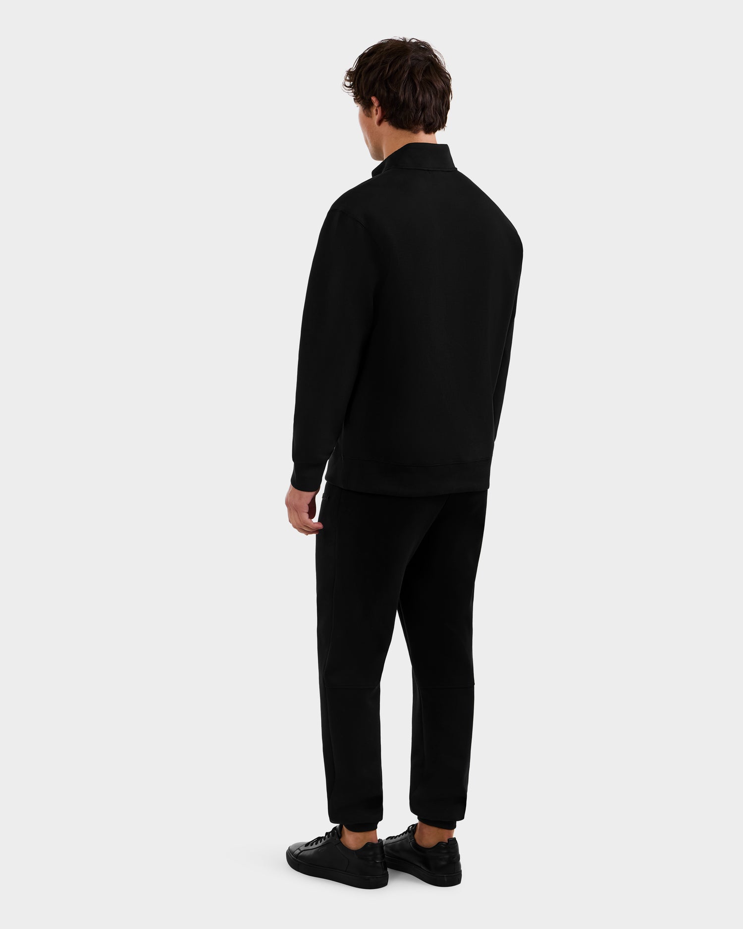 A person stands facing away, wearing the Rudsak FREMONT - 9525000 Black M sweater with black pants and shoes against a plain light gray background.