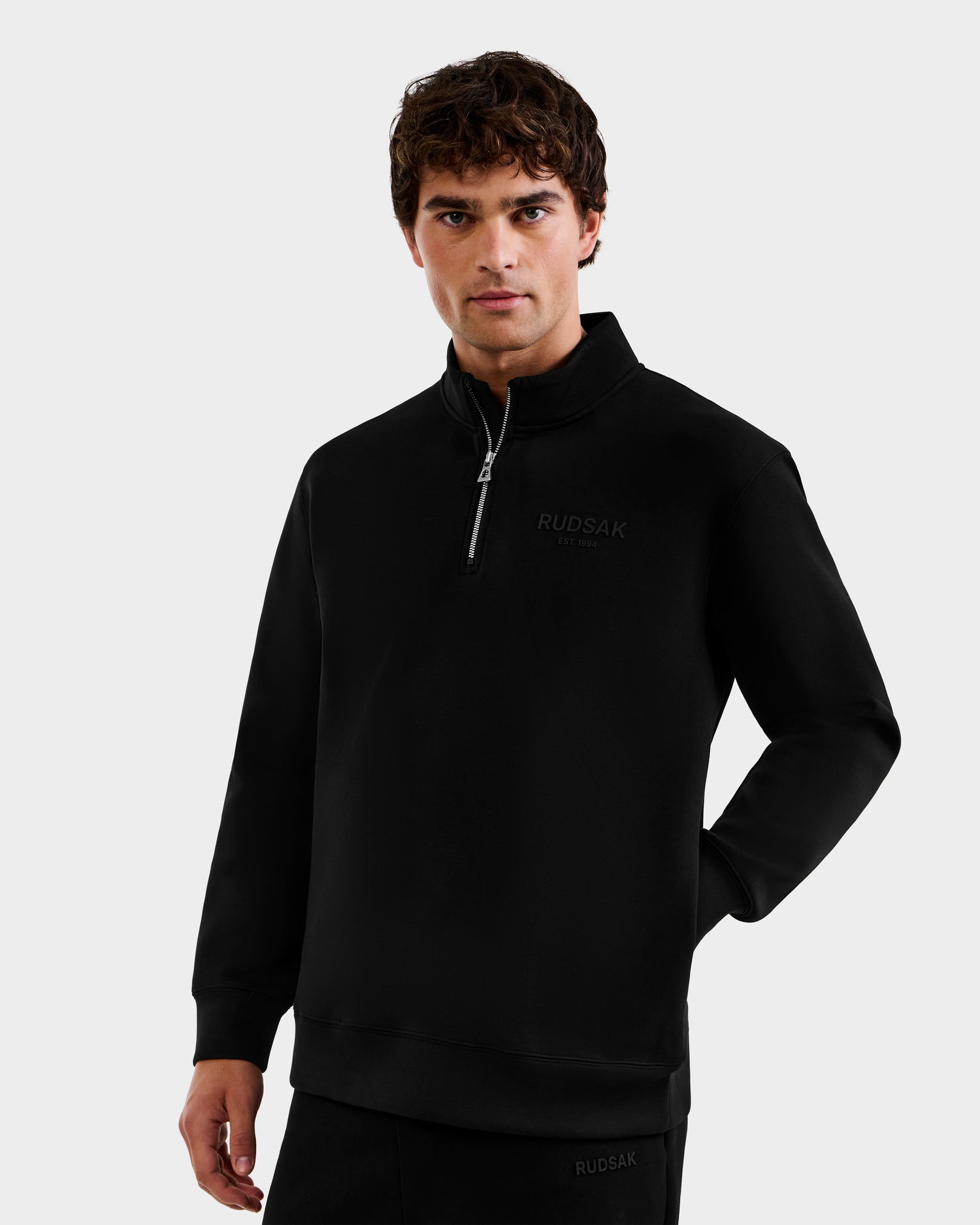 A man wears the Rudsak FREMONT - 9525000 quarter-zip pullover in black (size M), featuring chest text, standing against a plain light gray background with one hand in his pocket and the other by his side.