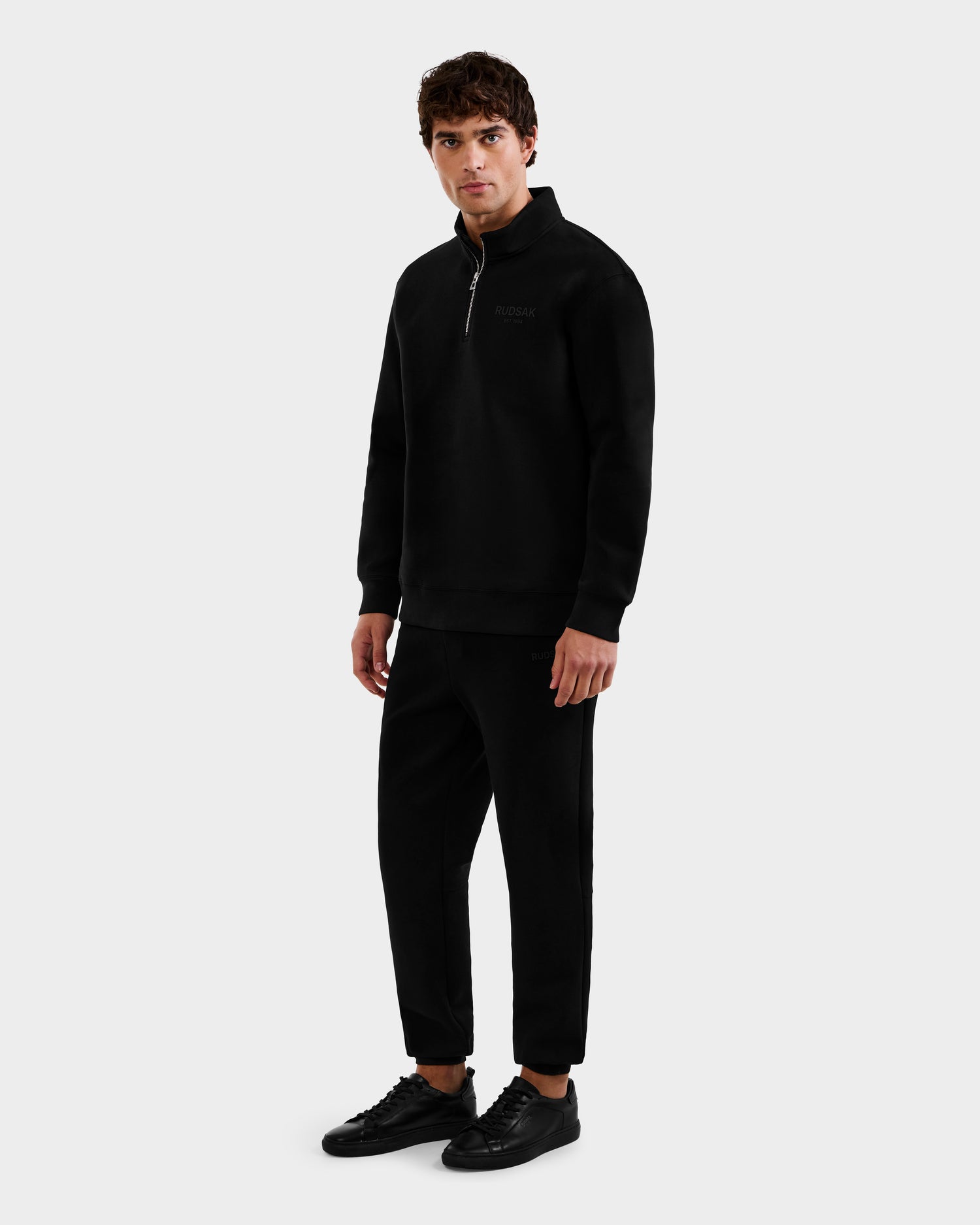 A man stands against a light gray background wearing the Rudsak FREMONT - 9525000 Black M mock neck sweater, paired with black pants and sneakers. He faces slightly right with a neutral expression.
