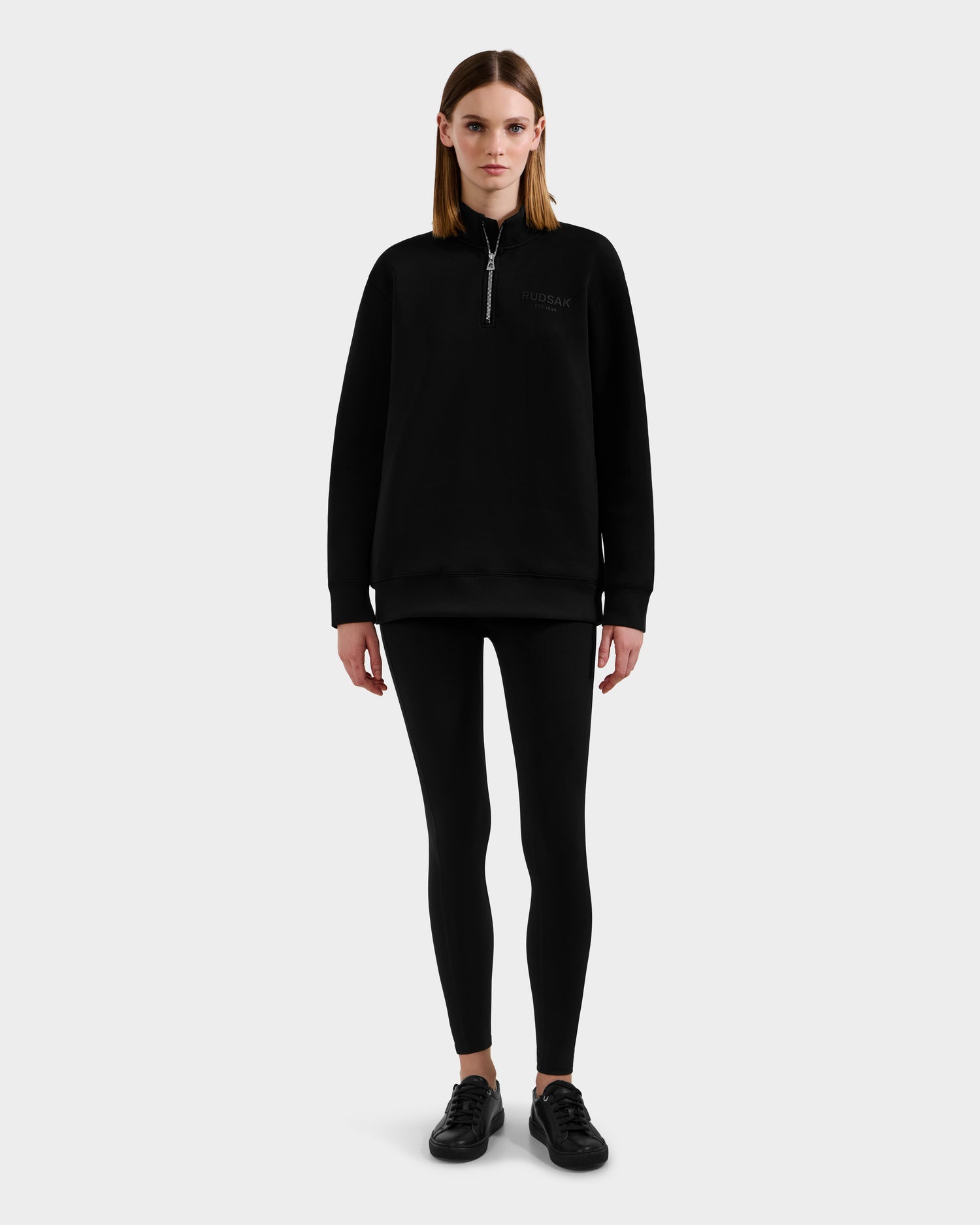 A woman faces forward, wearing the Rudsak FREMONT - 9525000 Black W zip-up sweatshirt with black leggings and sneakers, standing against a plain light gray background.
