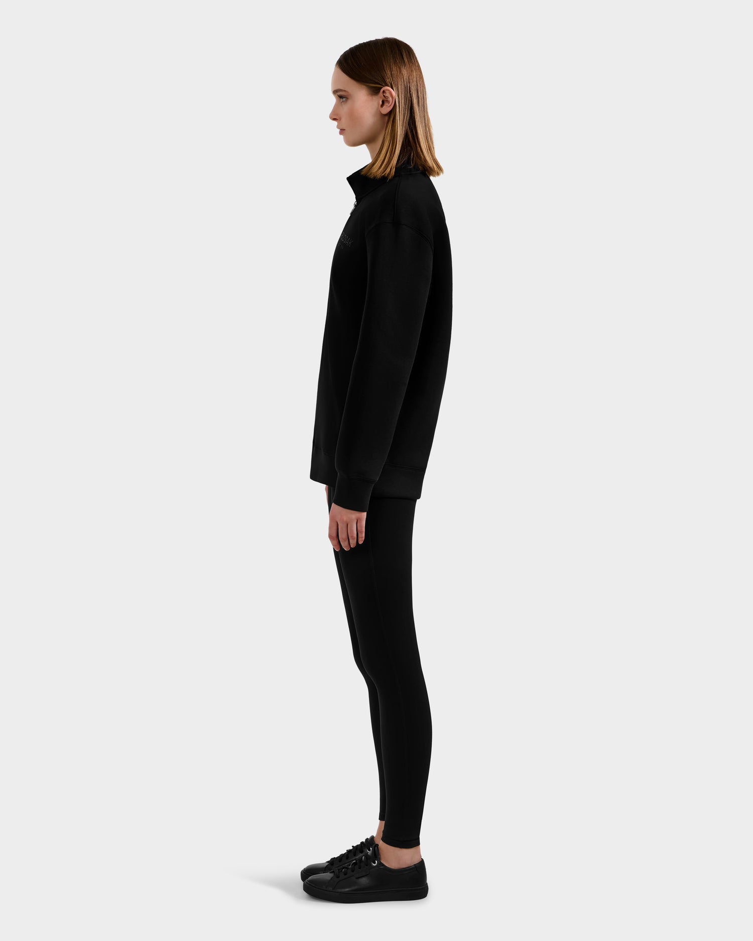A person stands in profile against a plain light background, wearing Rudsak's FREMONT - 9525000 Black W sweater, black leggings, and black sneakers.