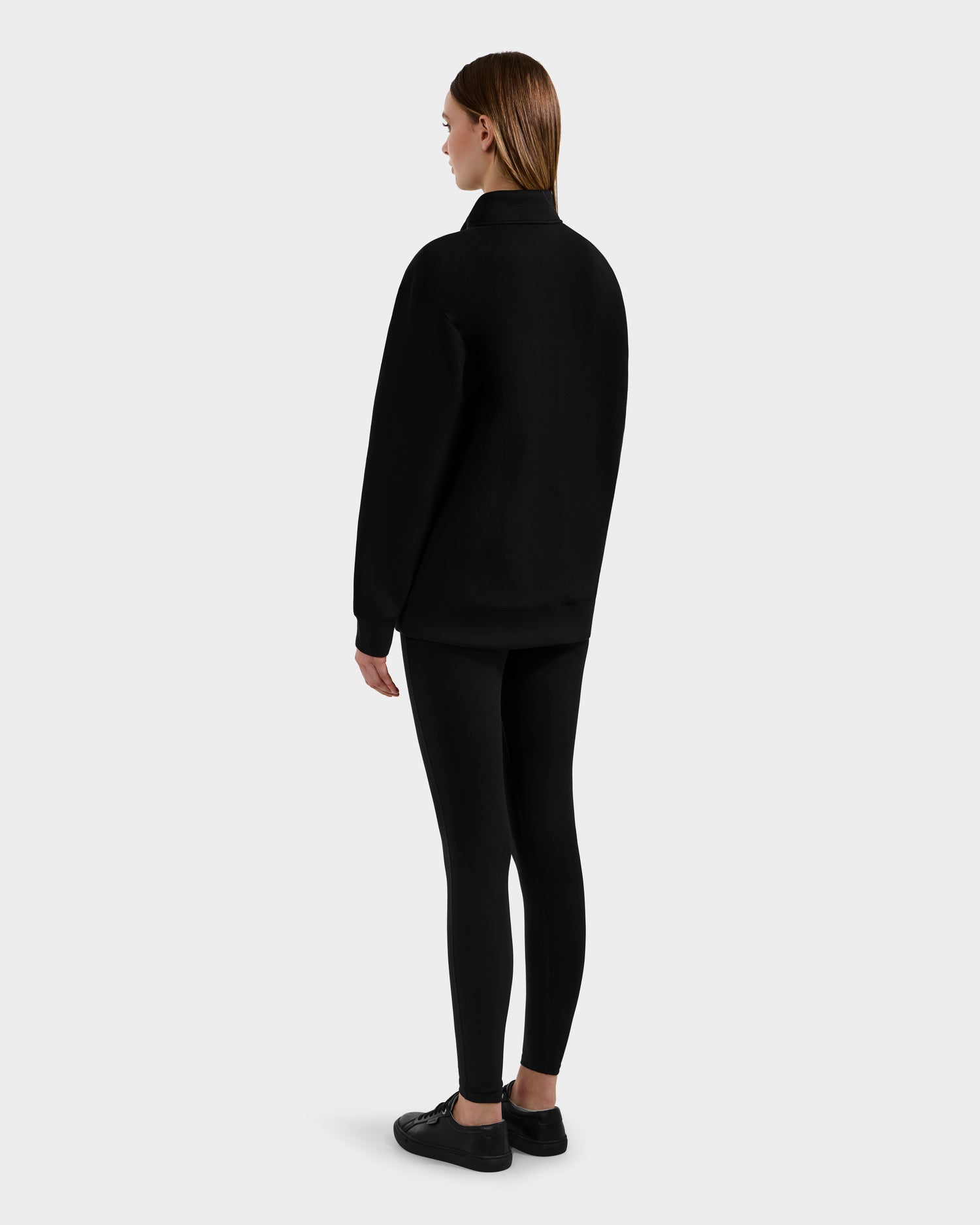 A woman with straight brown hair faces away, wearing the Rudsak FREMONT - 9525000 Black W mock neck cotton sweater, paired with black leggings and sneakers against a plain light gray background.