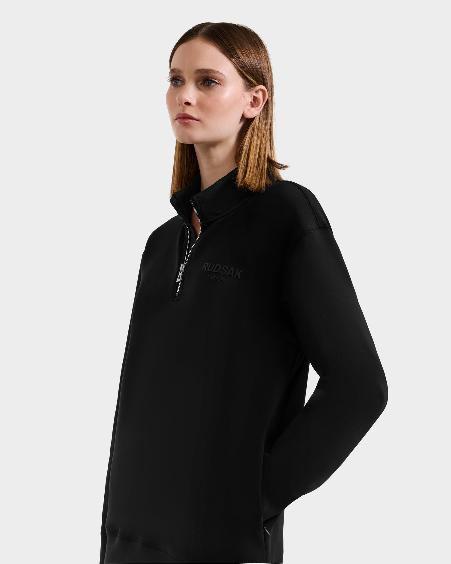 A woman with straight, shoulder-length brown hair wears the Rudsak FREMONT - 9525000 Black W premium cotton blend zip-collar sweatshirt with chest logo, standing against a light background and looking to the side.