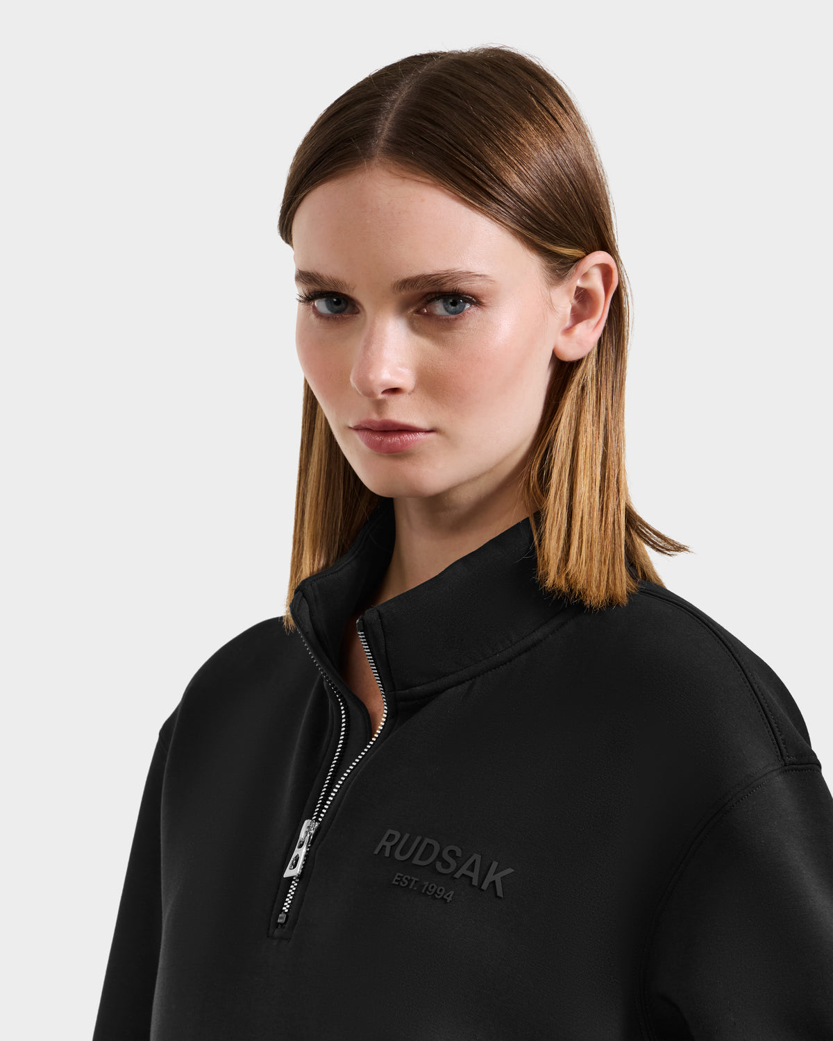 A woman with straight light brown hair wears the Rudsak FREMONT - 9525000 Black W zip-up top with "RUDSAK EST. 1994" embossed on the front, standing against a plain light background.
