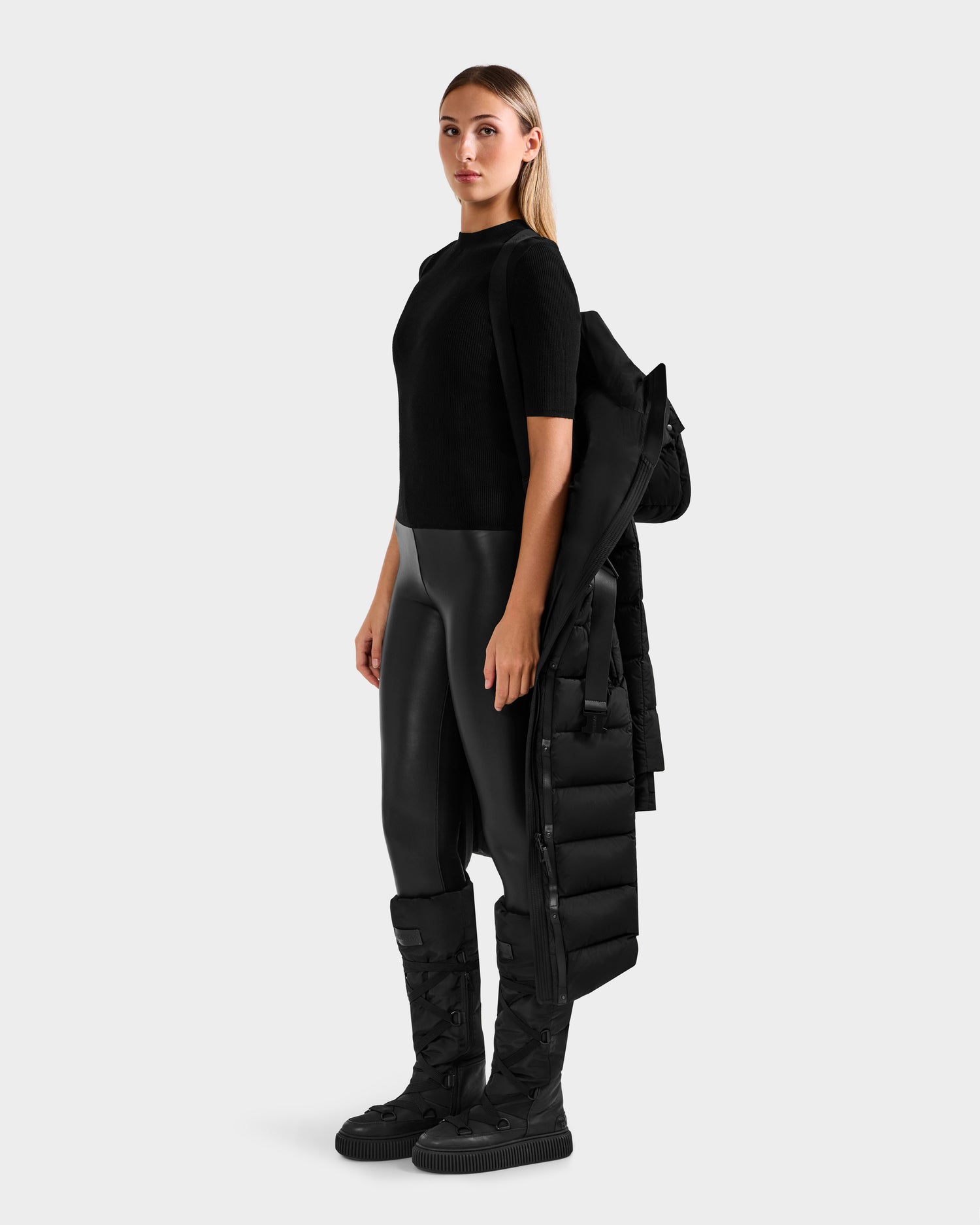 A woman stands facing forward, wearing the Rudsak FRIDA M-L - L125608 Black quilted down coat draped over one arm against a plain light gray background.