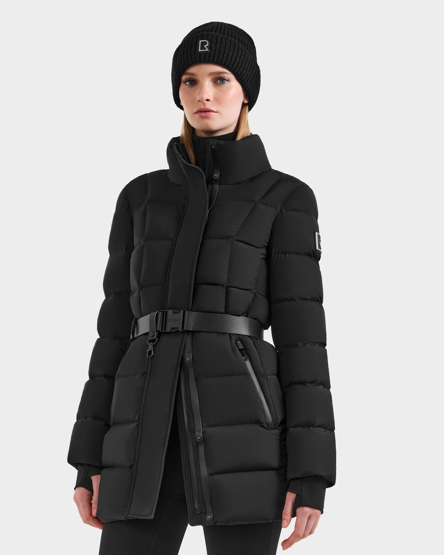 A woman wears the Rudsak FRIDA M-M - L125607 Black quilted down puffer coat with belted waist and detachable hood, plus a matching black knit beanie, standing against a plain light background—your versatile winter essential.