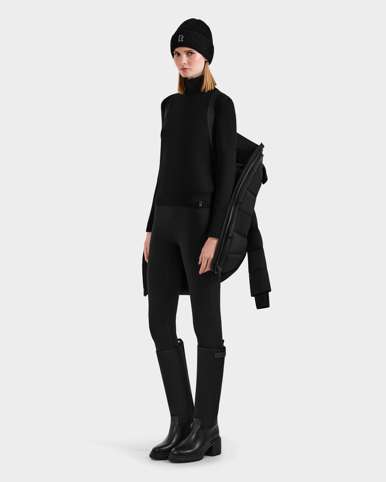 A woman in all-black attire stands confidently against a plain light background, featuring the Rudsak FRIDA M-M - L125607 Black quilted down puffer with detachable hood draped over her shoulder.