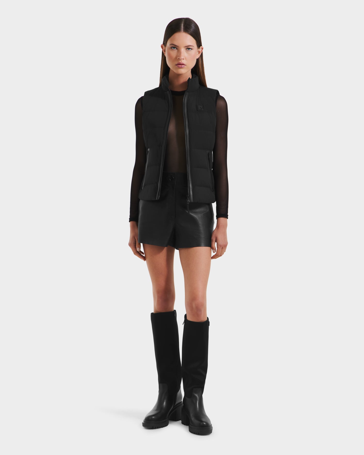 A woman faces forward against a plain white background, wearing the Rudsak GABRIELA - 8125014 Black lightweight puffer vest with vegan leather trims over a sheer black top, black leather shorts, and knee-high black boots.