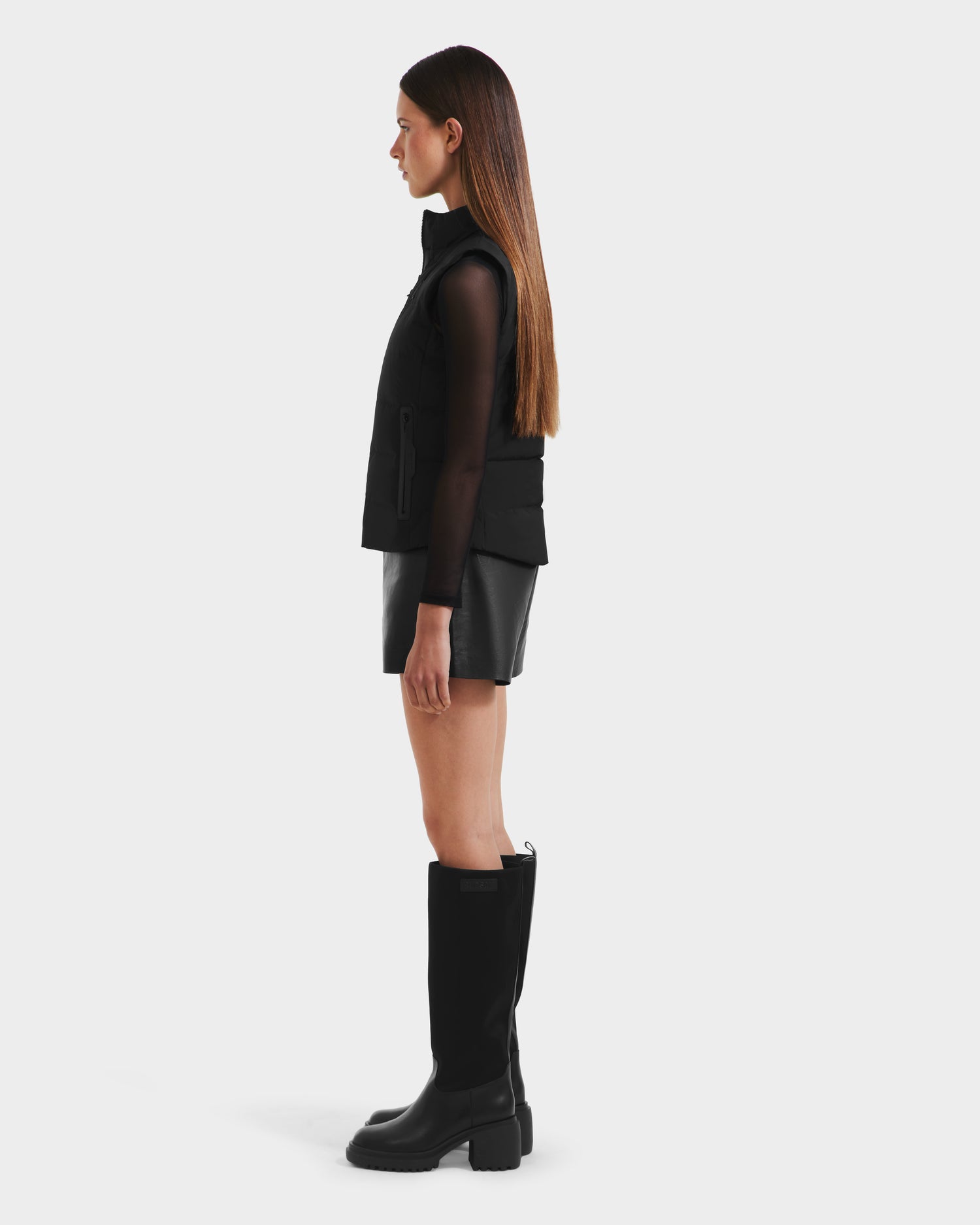 A woman in profile wears the Rudsak GABRIELA - 8125014 puffer vest with vegan leather trims, styled with a sheer long-sleeve top, black leather skirt, and knee-high boots against a plain white background.