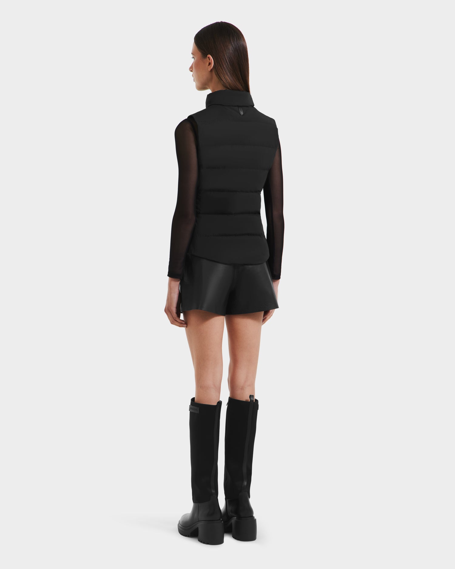 A woman stands facing away, wearing the Rudsak GABRIELA - 8125014 Black womens down puffer vest with vegan leather trims, paired with a black sheer long-sleeve top, black leather shorts, and knee-high black boots against a plain light background.
