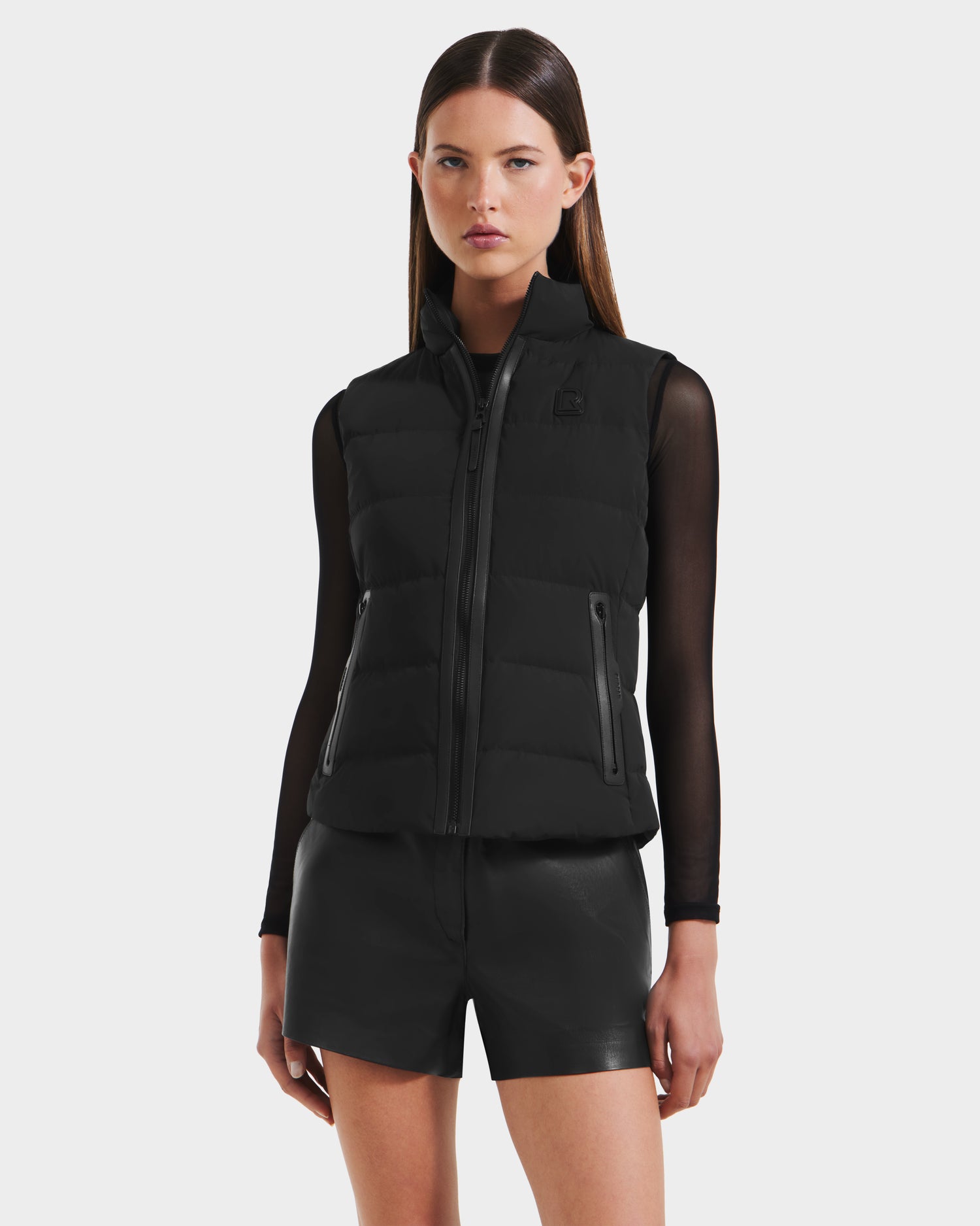 A woman with straight brown hair models the Rudsak GABRIELA - 8125014 Black lightweight puffer vest with vegan leather trims over a sheer black long-sleeve top and black leather shorts, set against a plain light background.