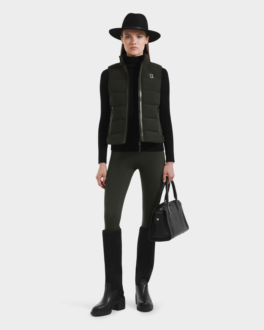 A person wearing a black hat, black turtleneck, and the Rudsak GABRIELA - 8125014 Down Puffer Vest in Dark Olive with vegan leather trims, paired with dark pants, black tall boots, and holding a black handbag, stands against a plain white background.