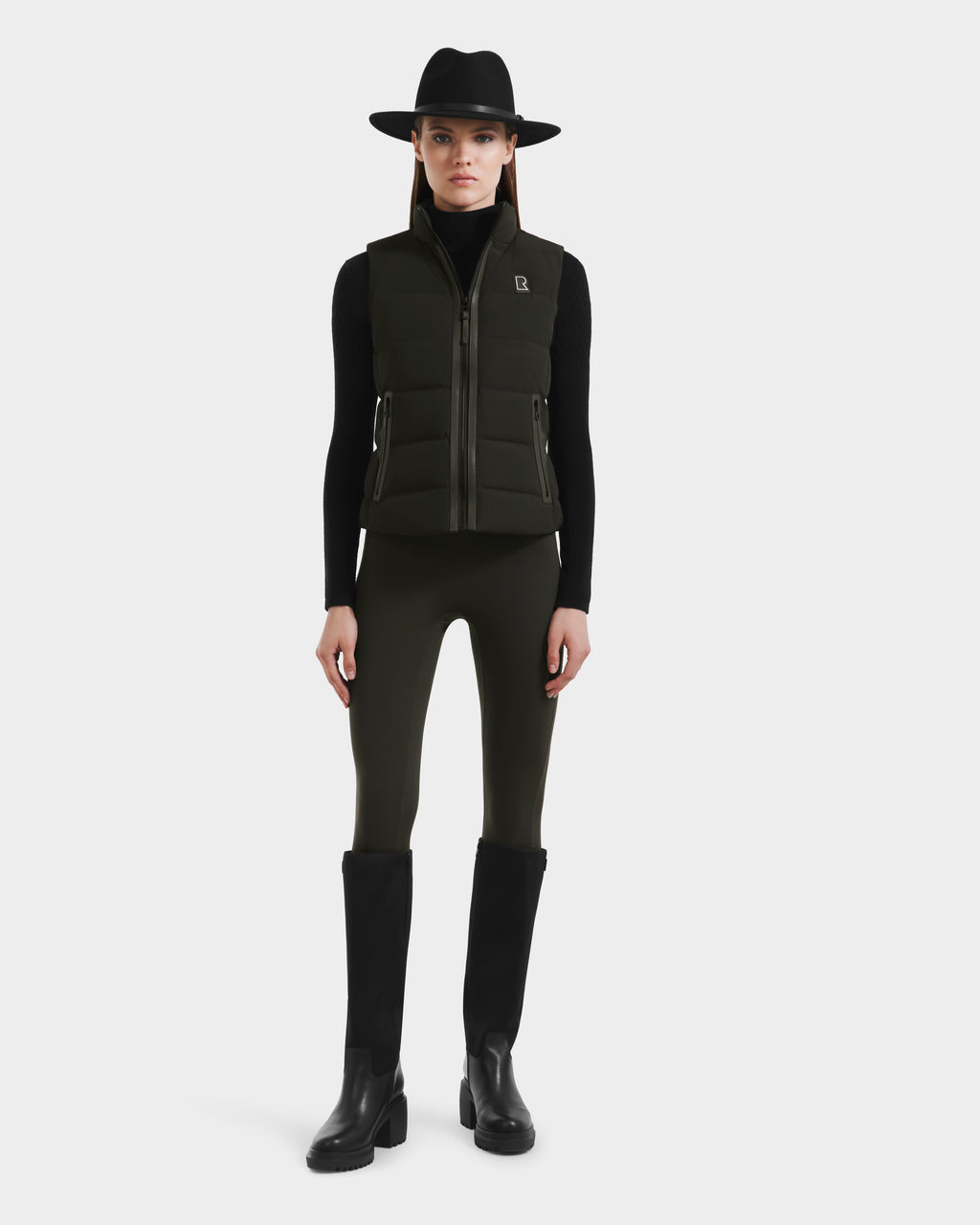 A woman wearing a black wide-brim hat, a black long-sleeve top, the Rudsak GABRIELA - 8125014 Dark Olive women’s down puffer vest with vegan leather trims, green leggings, and black knee-high boots stands facing forward against a plain white background.