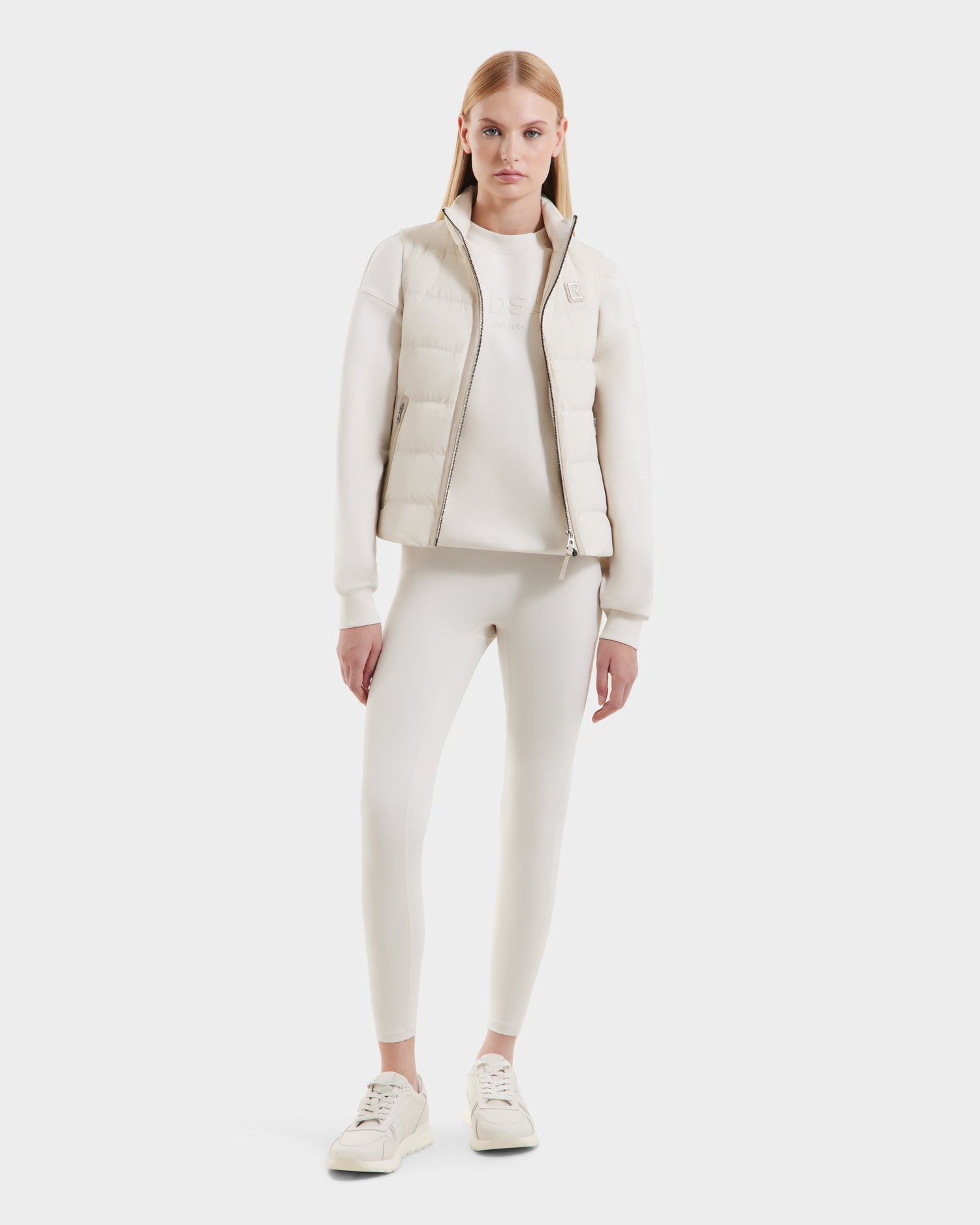 A woman faces forward against a plain white background, wearing the Rudsak GABRIELA – 8125014 Vanilla: a lightweight all-cream puffer vest with vegan leather trims, styled with a matching long-sleeve top, leggings, and sneakers.