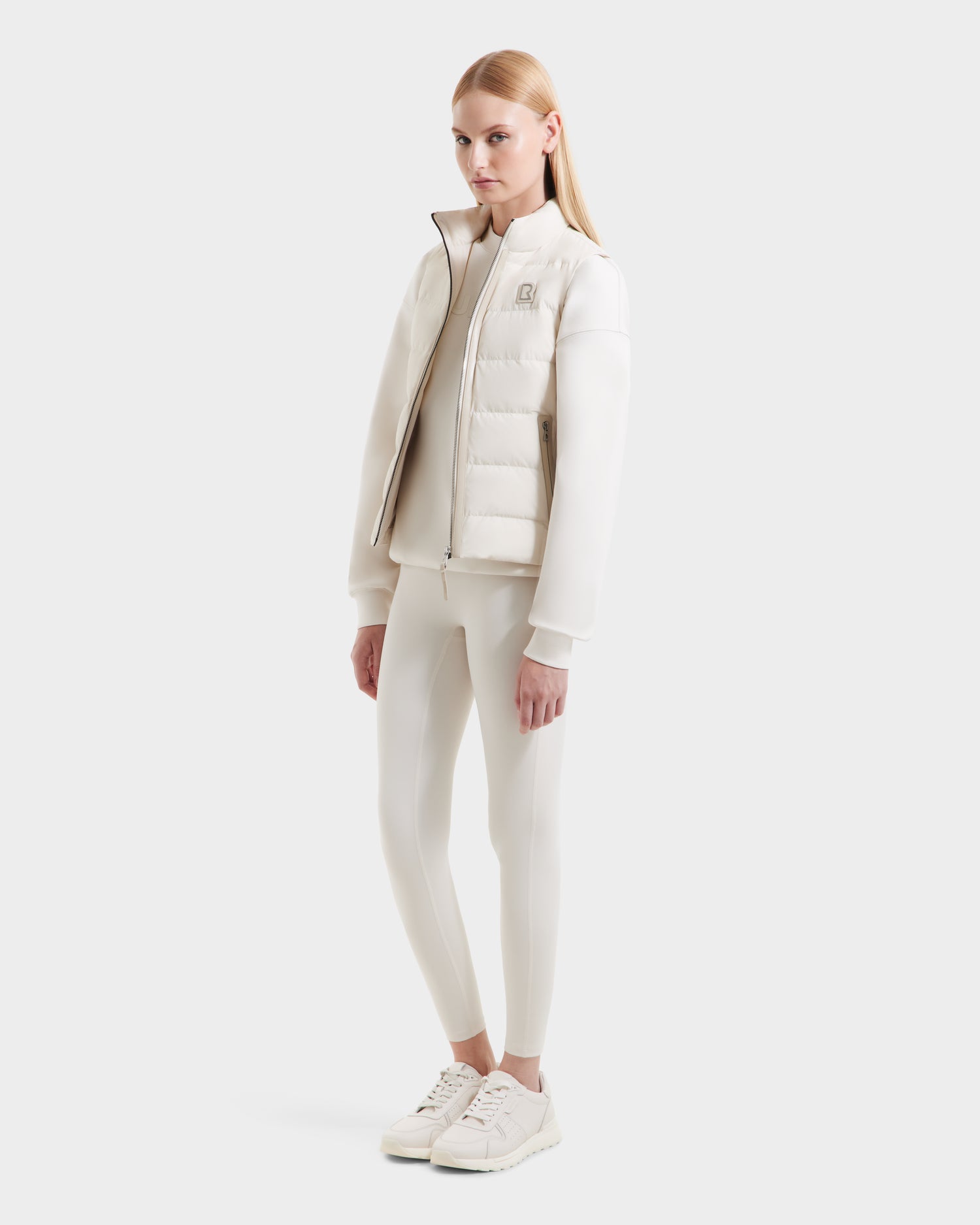 A woman with straight blonde hair wears the Rudsak GABRIELA - 8125014 Vanilla puffer jacket with vegan leather trims, leggings, and sneakers against a white background. She stands relaxed, facing slightly left with a neutral expression.
