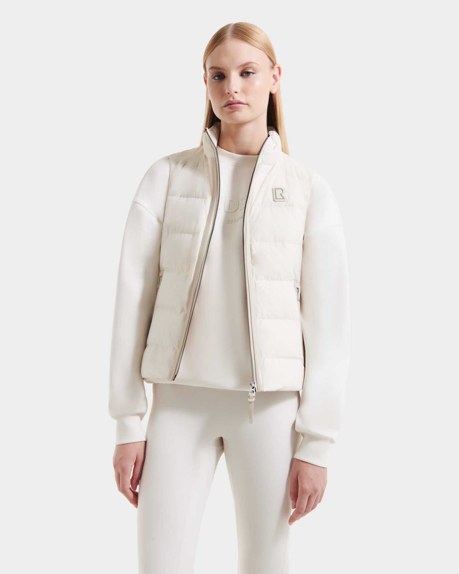 A woman with straight blonde hair models the Rudsak GABRIELA - 8125014 Vanilla women’s fitted down puffer vest with vegan leather trims over a matching long-sleeve top and pants against a plain white background.