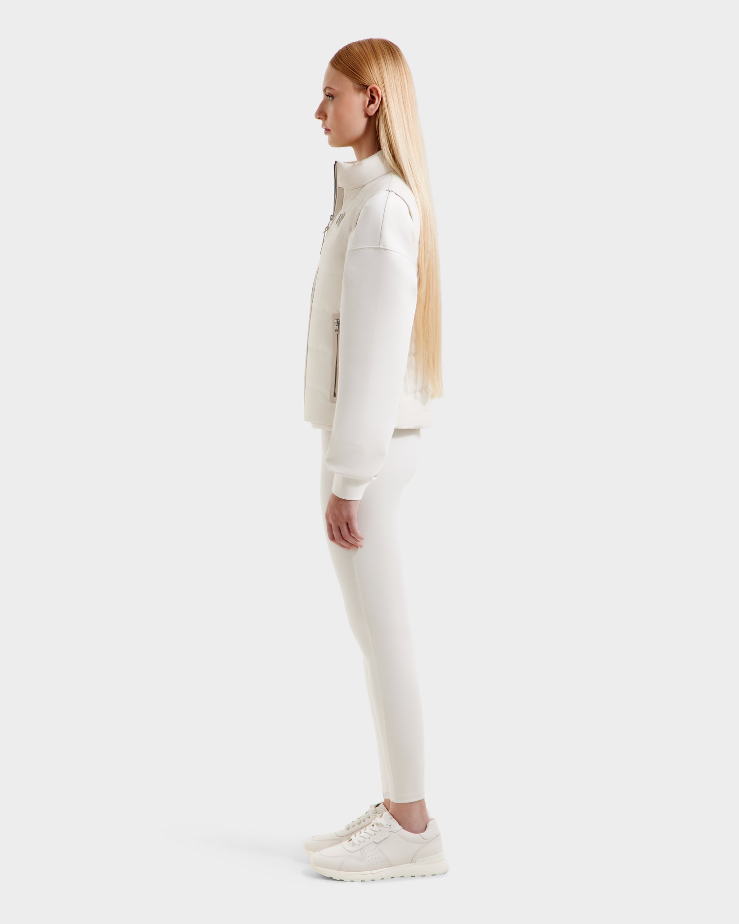 A woman with long straight blonde hair stands in profile, wearing Rudsak’s GABRIELA - 8125014 Vanilla lightweight down vest with vegan leather trim, paired with leggings and sneakers, against a plain white background.