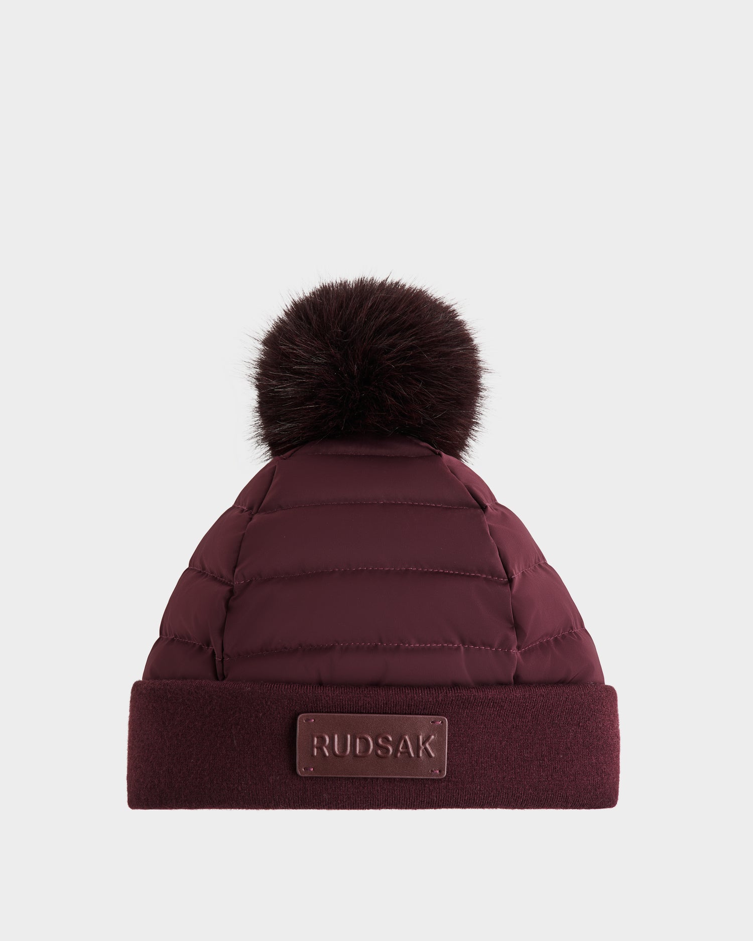 The GALA - 8424514 Bordeaux by Rudsak is a women's quilted down puffer winter hat in burgundy, featuring horizontal stitching, a dark faux fur pom-pom, a folded brim, and a Rudsak logo patch. Plain white background.
