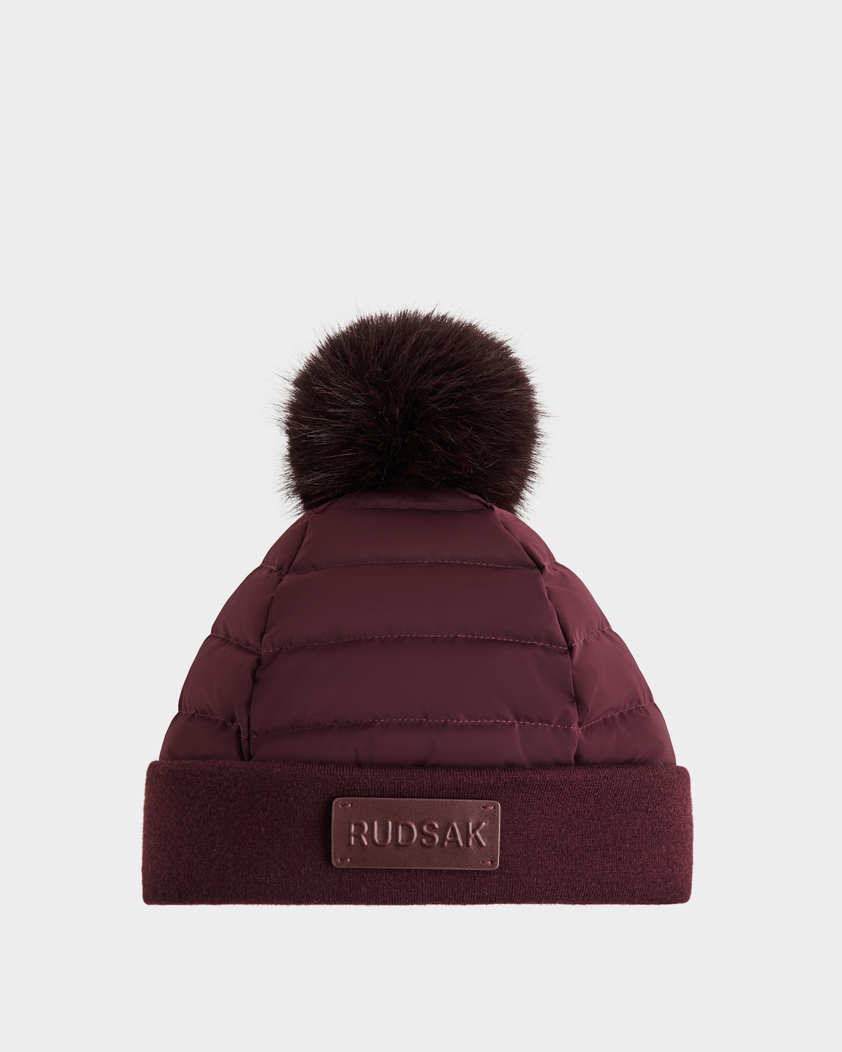 The GALA - 8424514 Bordeaux by Rudsak is a women's quilted down puffer winter hat in burgundy, featuring horizontal stitching, a dark faux fur pom-pom, a folded brim, and a Rudsak logo patch. Plain white background.