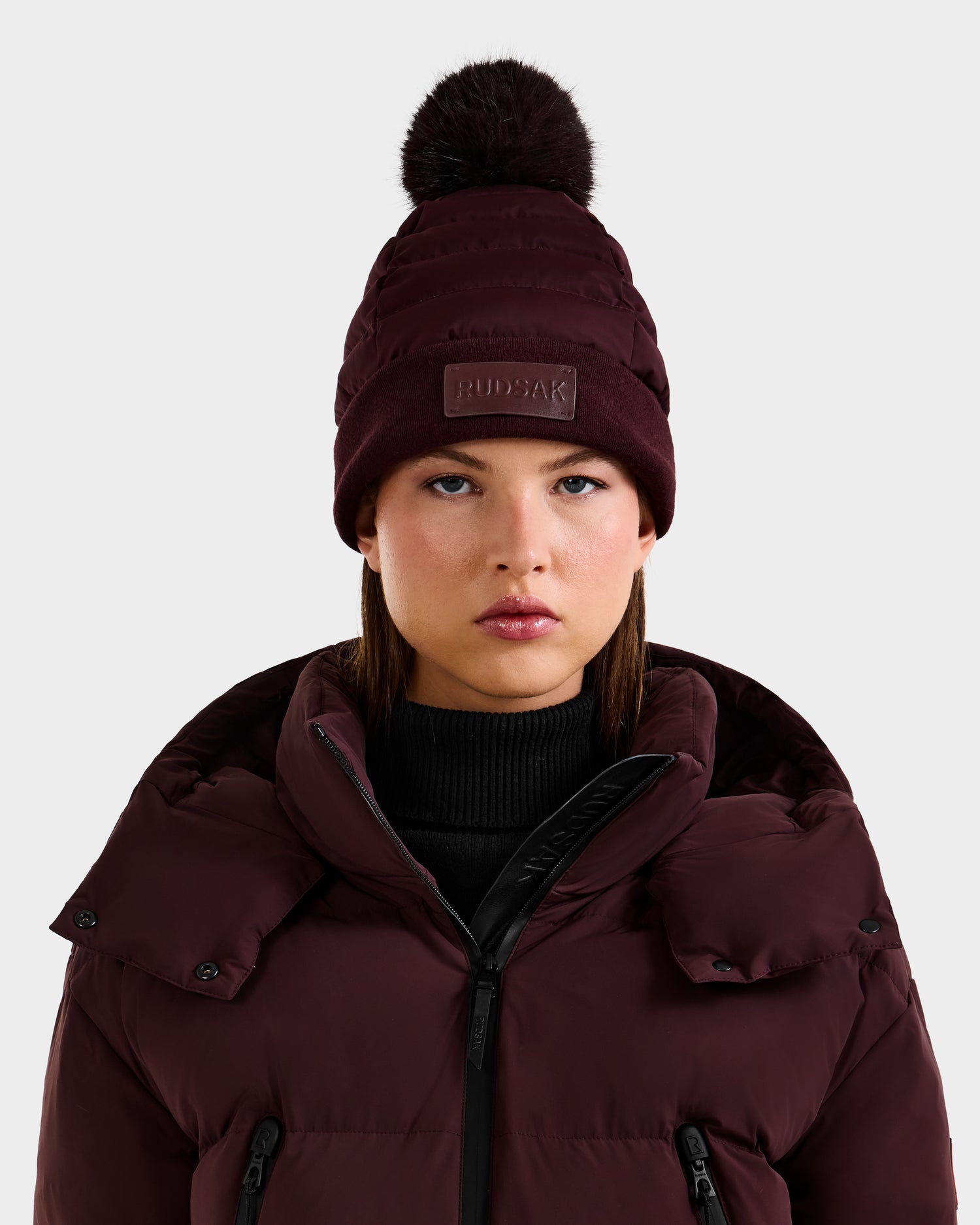A woman wears the Rudsak GALA - 8424514 Bordeaux quilted down puffer hat with a removable faux fur pom pom and a matching burgundy puffer jacket. She stands against a plain light background, her expression neutral.