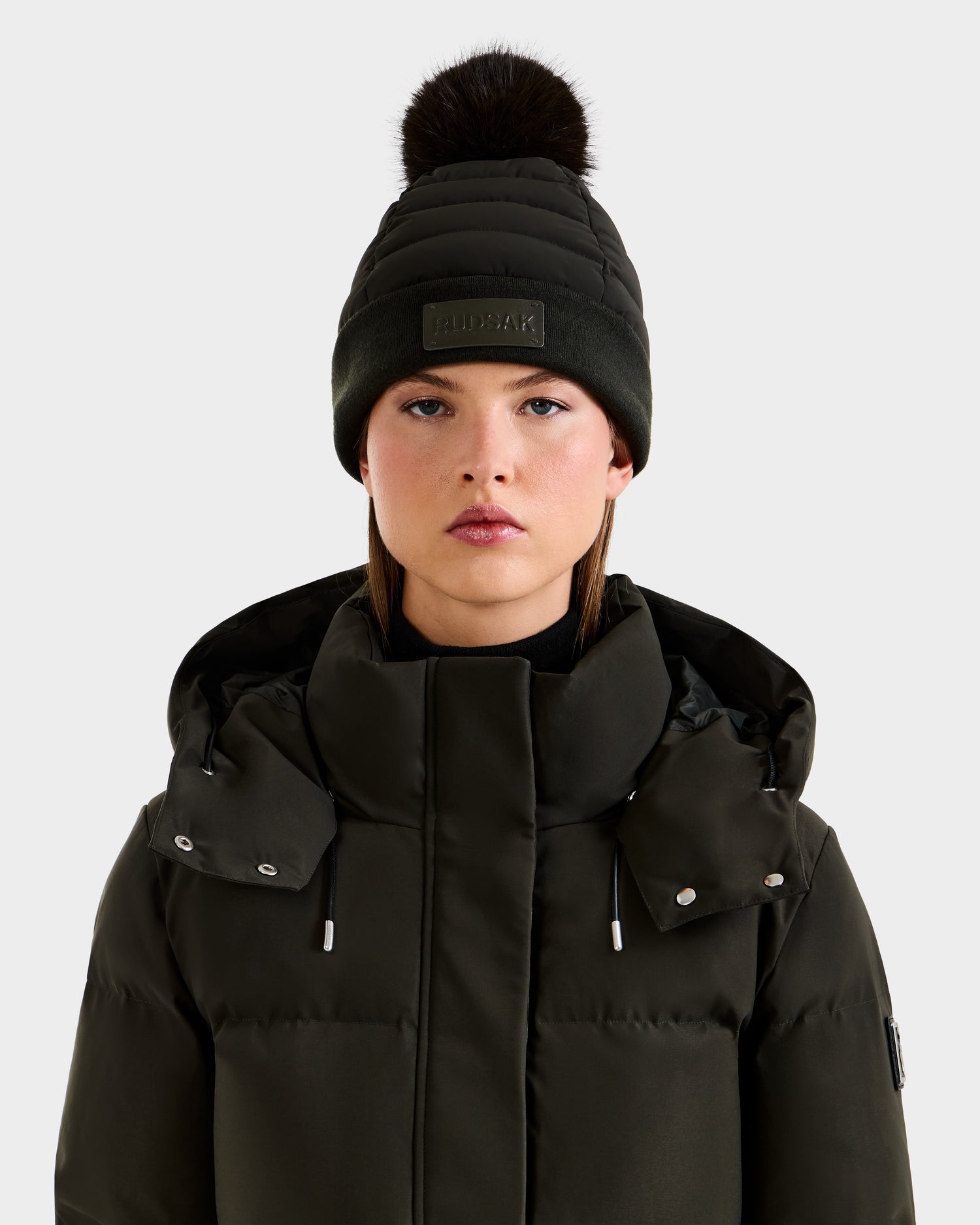 A woman wears the Rudsak GALA - 8424514 Dark Olive puffer jacket with a hood and a matching women's quilted down puffer hat with a faux fur pom pom, looking straight ahead against a light background.