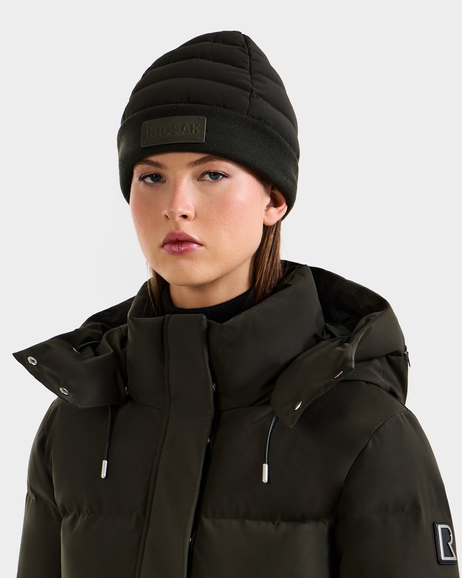 A woman wears the Rudsak GALA - 8424514 Dark Olive puffer jacket with a high collar and a women's winter hat, standing against a plain light background and looking directly at the camera with a neutral expression.