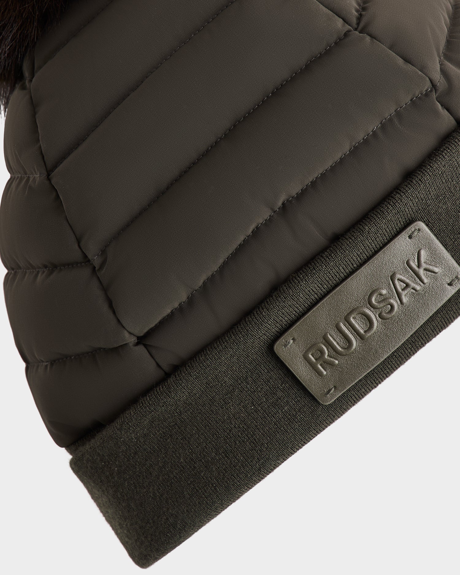 Close-up of the Rudsak GALA - 8424514 women’s dark olive quilted down puffer hat with a ribbed cuff, featuring a rectangular Rudsak patch and topped with a removable faux fur pom pom.