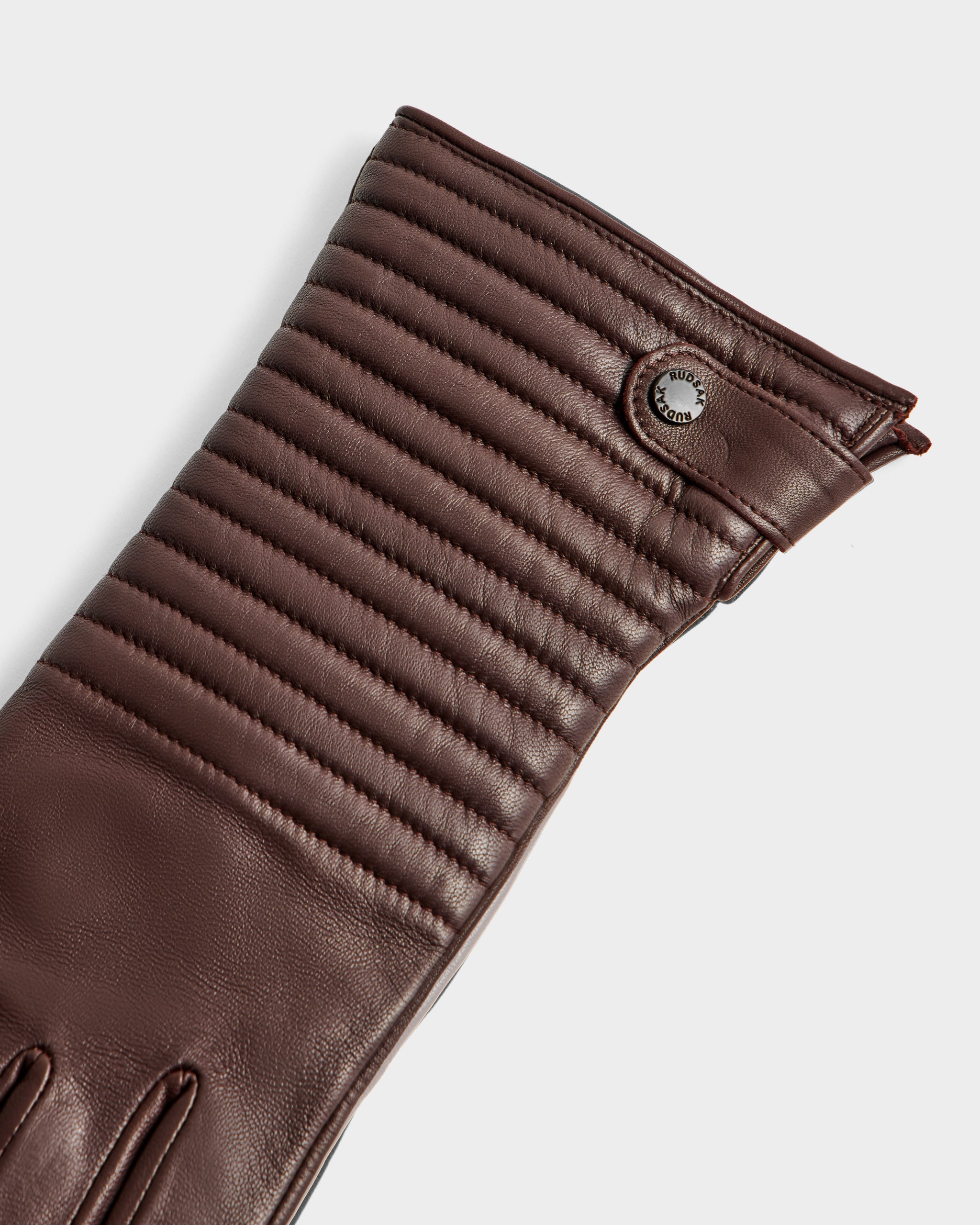 A close-up of the GEMMA - 8425008 Espresso brown leather glove by Rudsak, featuring stitched horizontal ridges on the cuff and a metal button detail—an elegant winter accessory, cashmere-lined for added comfort.
