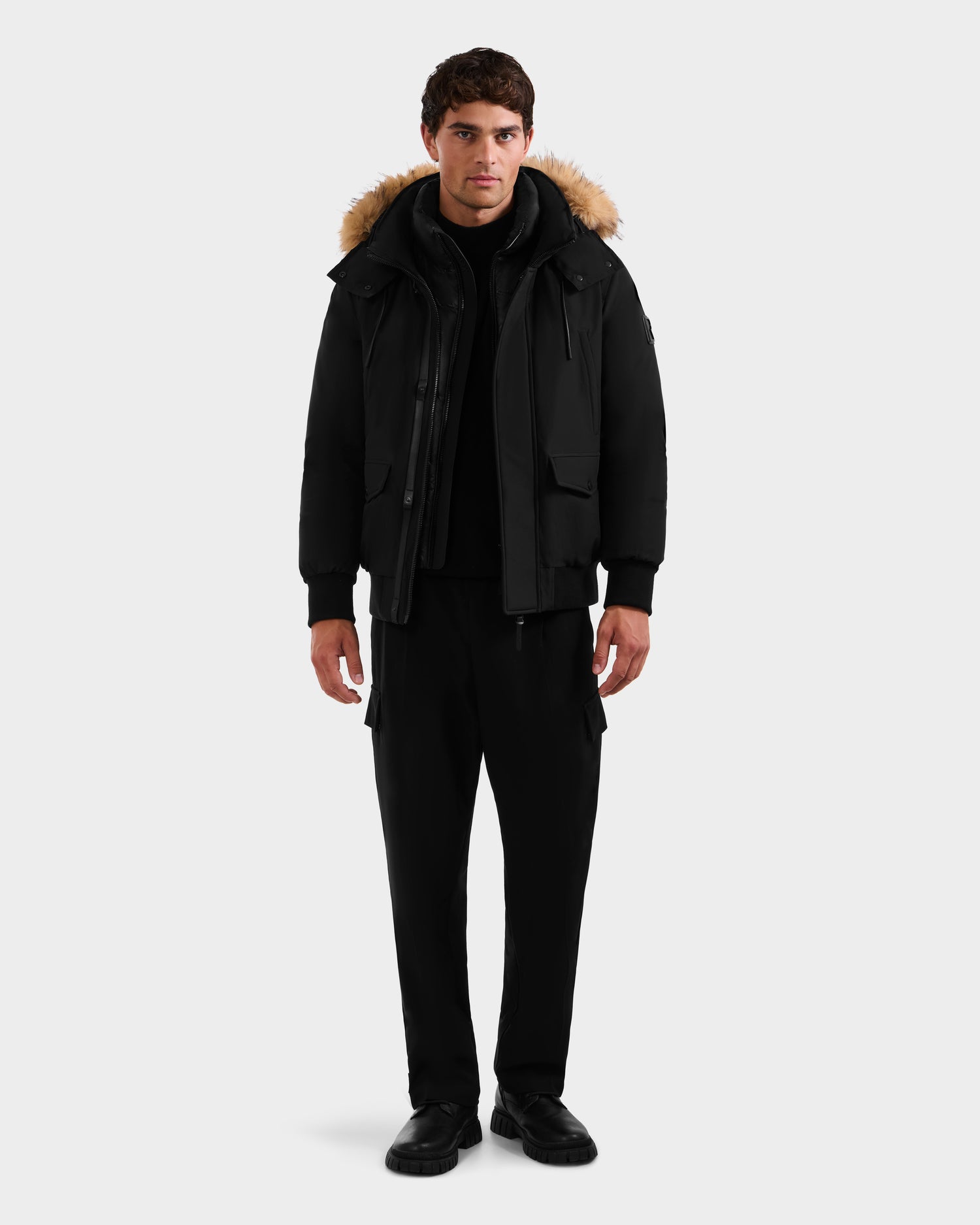 A man wears the Rudsak GEORGE - M125511 Black men's winter down bomber with a fur-trimmed removable hood, black pants, and black shoes against a plain light gray background.