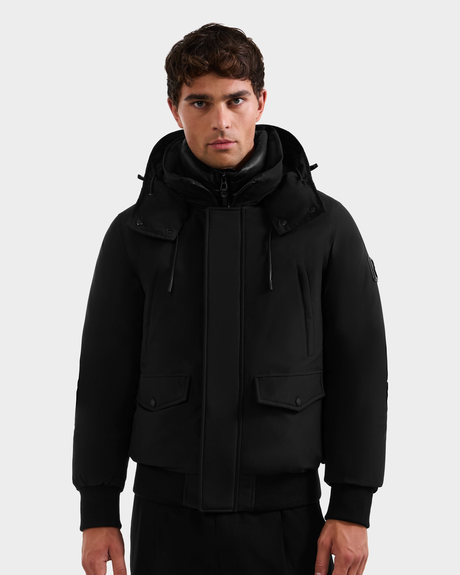A young man with short, wavy hair wears the Rudsak GEORGE - M125511 Black men’s winter down bomber with a high collar, front flap pockets, ribbed cuffs, and a removable hood, standing against a plain light gray background.