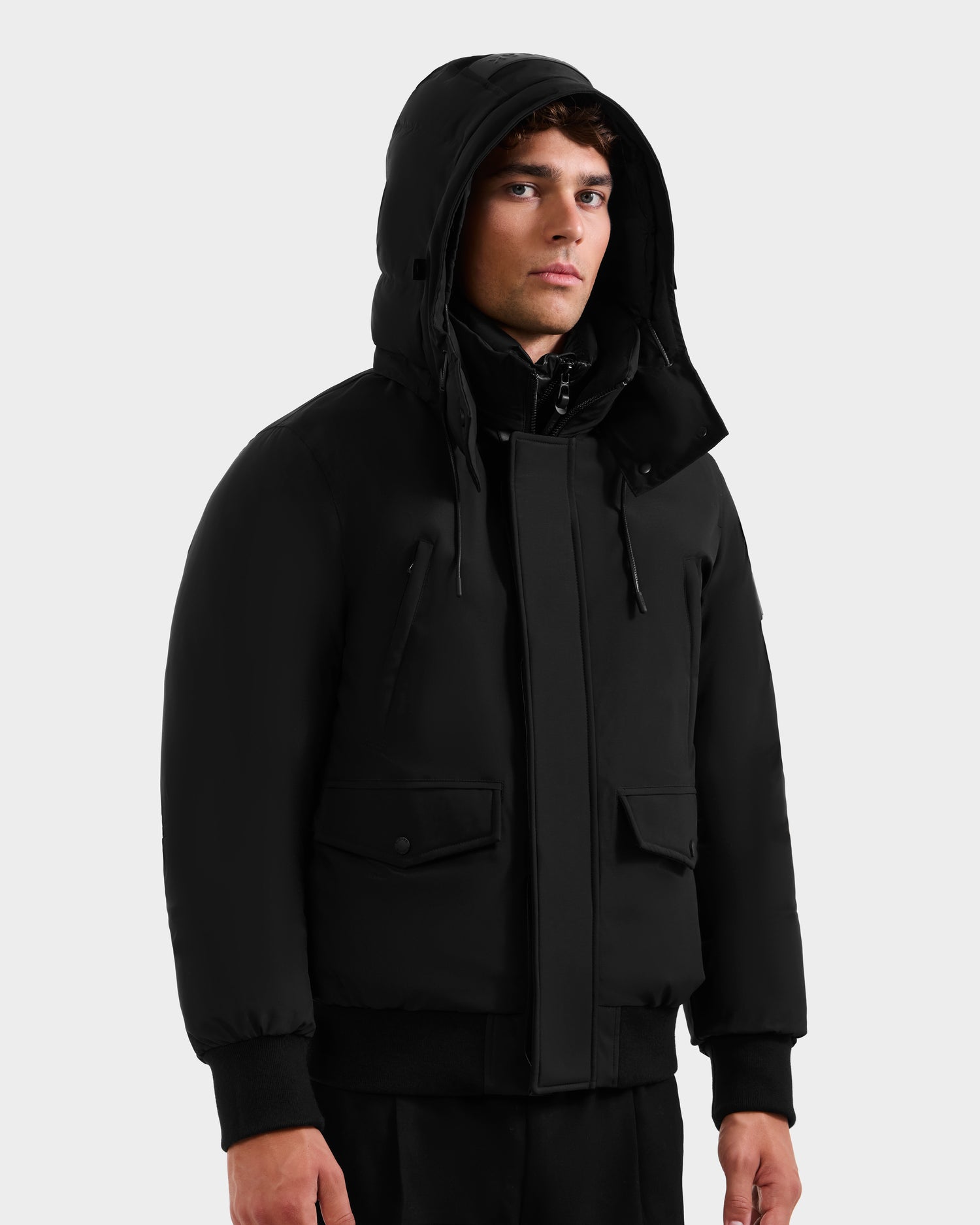 A man wearing the Rudsak GEORGE - M125511 Black men’s winter down bomber with flap pockets and a removable hood stands against a plain light gray background.