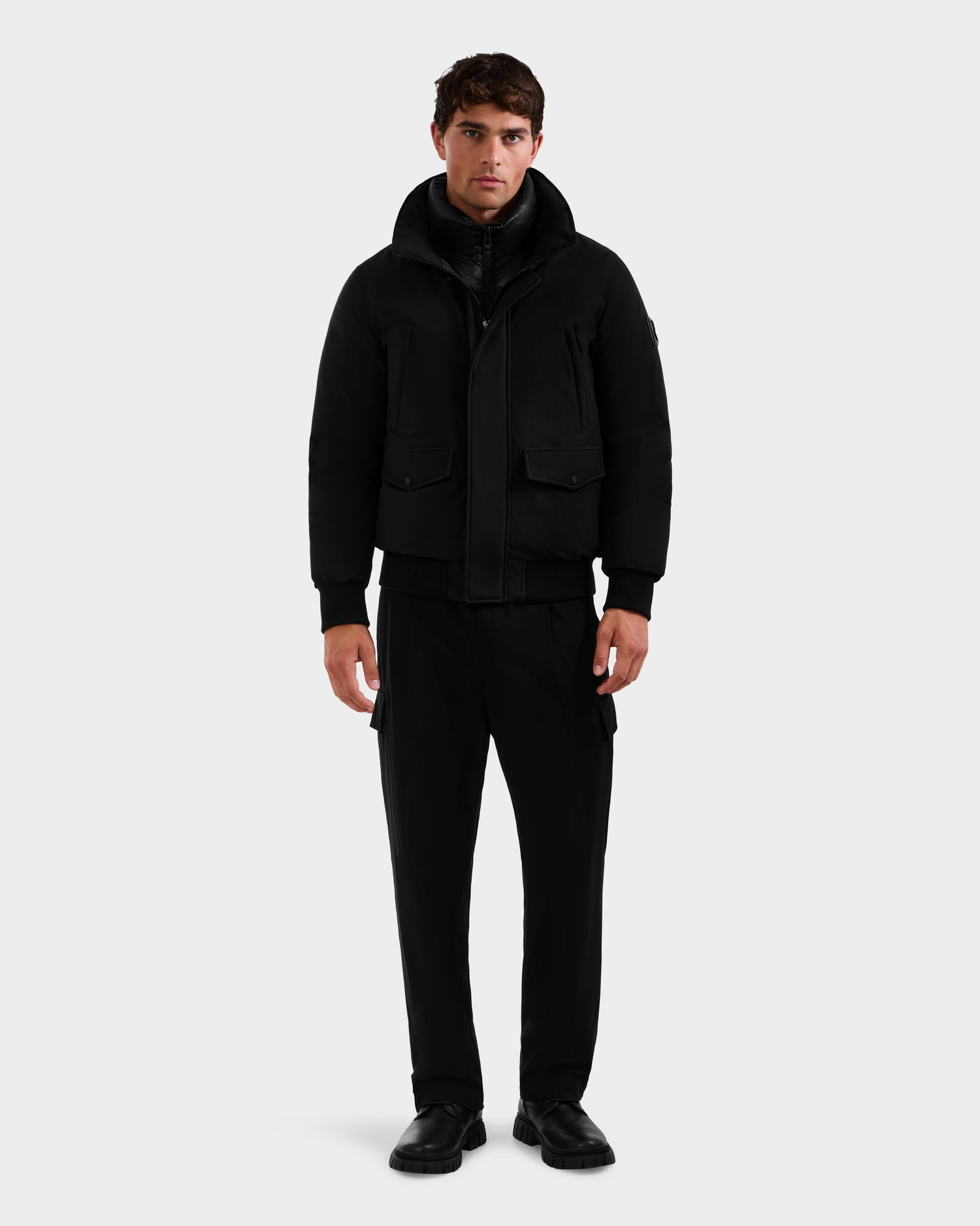 A man with short dark hair and a neutral expression faces forward, wearing the Rudsak GEORGE - M125511 Black men’s winter down bomber, paired with black pants and shoes, against a plain light gray background.