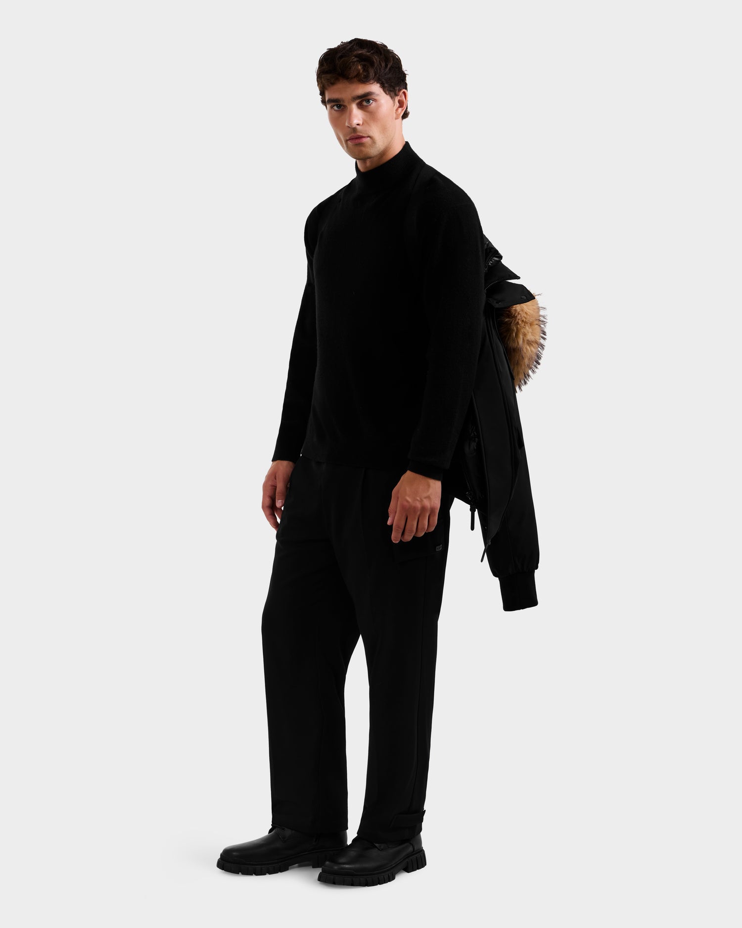 A man in an all-black outfit holds the Rudsak GEORGE - M125511 Black men's winter down bomber with removable hood and fur lining, casually draped over his shoulder against a plain background.