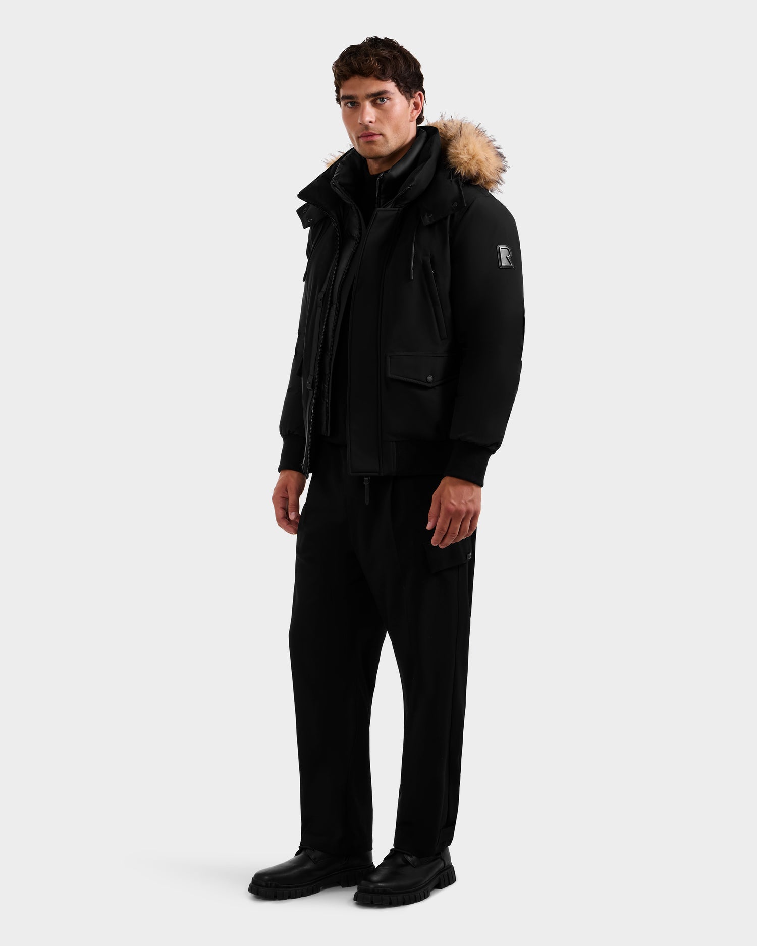 A man stands against a plain background wearing the Rudsak GEORGE - M125511 Black men’s winter down bomber with a fur-lined, removable hood, black pants, and black shoes. He faces slightly left, expression neutral.