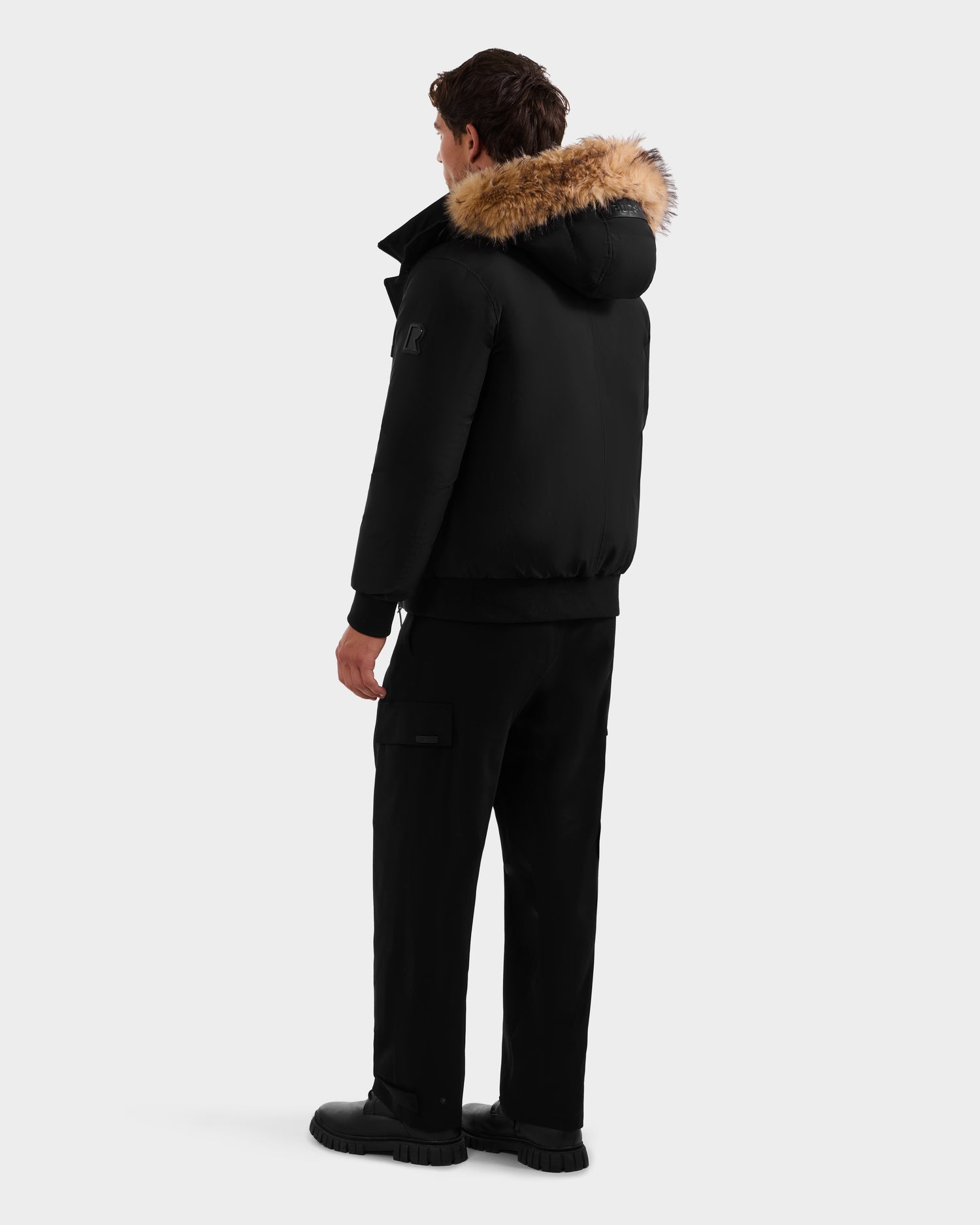 A person stands facing away, wearing the Rudsak GEORGE - M125511 Black men’s down bomber with brown fur trim and removable hood, paired with black pants and shoes, against a plain light background.