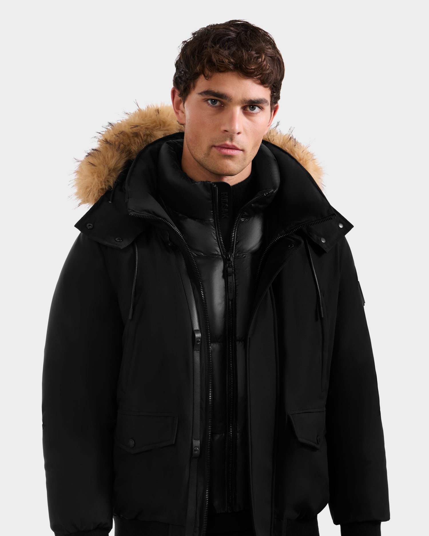 A man with short brown hair wears the Rudsak GEORGE - M125511 Black, a premium men’s winter down bomber with a black fur-lined removable hood and zippered inner layer, standing against a plain light gray background.