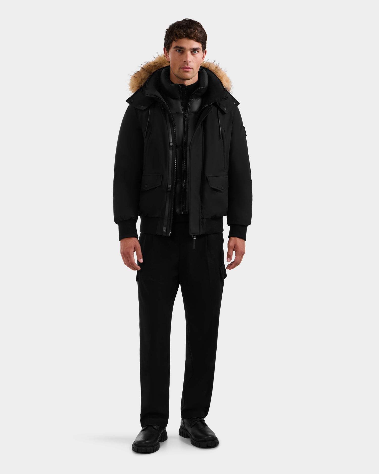 A man stands facing forward wearing the Rudsak GEORGE - M125511 Black men’s winter down bomber with a fur-lined, removable hood, plus black shirt, pants, and shoes, against a plain light background.