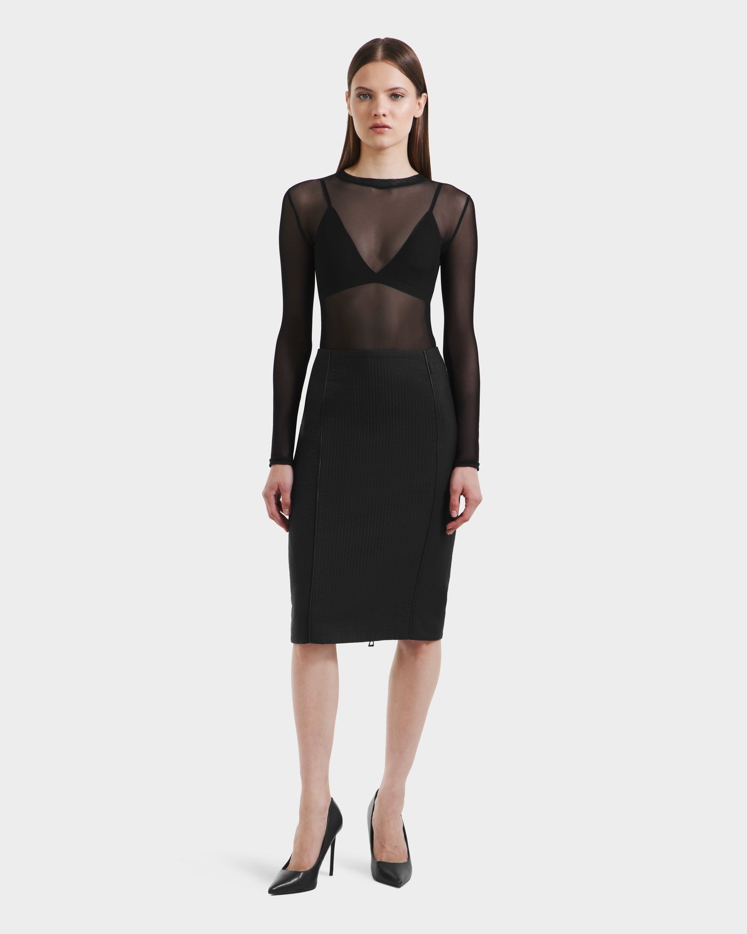 A woman faces forward in a Rudsak GLENDORA - 8525563 Black sheer long-sleeve top over a black bralette, paired with a fitted skirt and high heels, set against a plain white background.