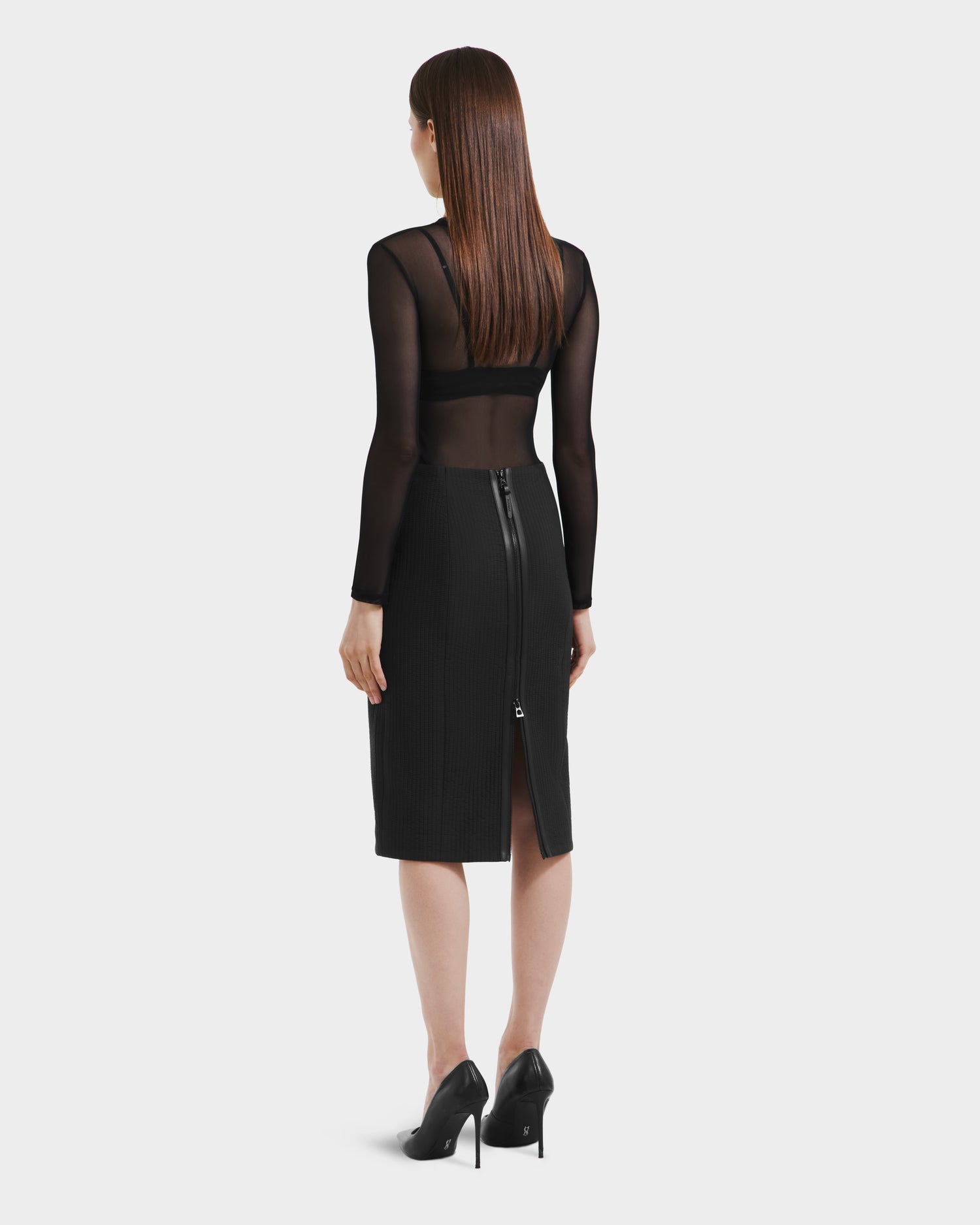 A woman with straight brown hair wears the Rudsak GLENDORA - 8525563 Black vegan leather fitted midi skirt with a visible back zipper and slit, styled with a sheer black top and black high heels, facing away from the camera.