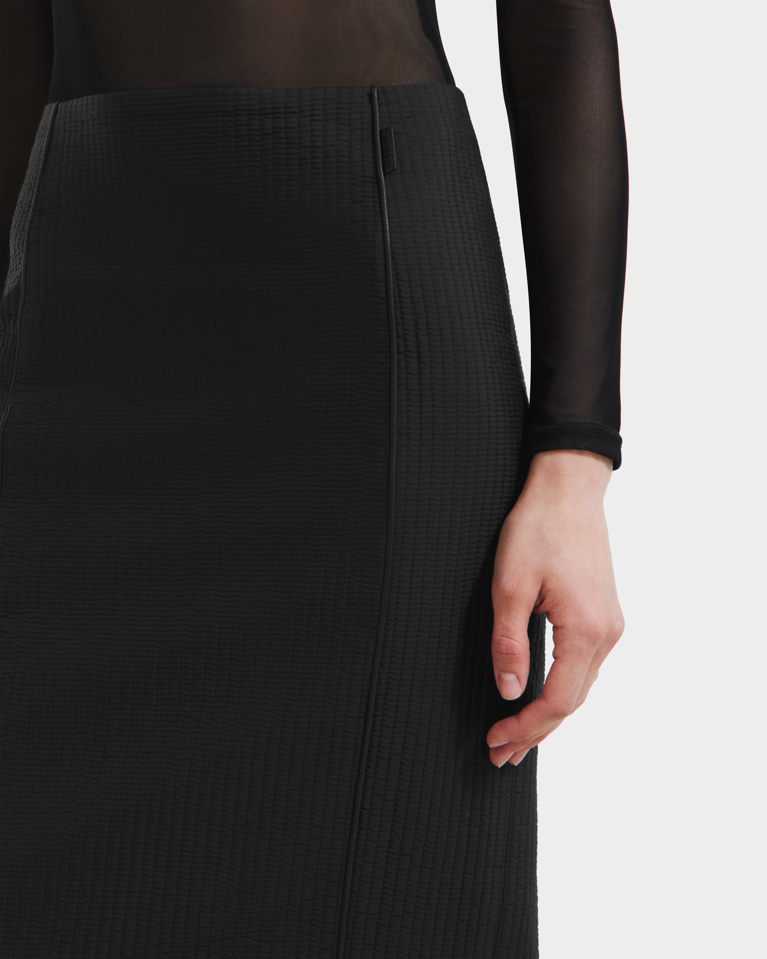 A model wears the Rudsak GLENDORA - 8525563 Black vegan leather fitted skirt with a sheer black long-sleeve top, shown waist to mid-thigh, right arm by their side.