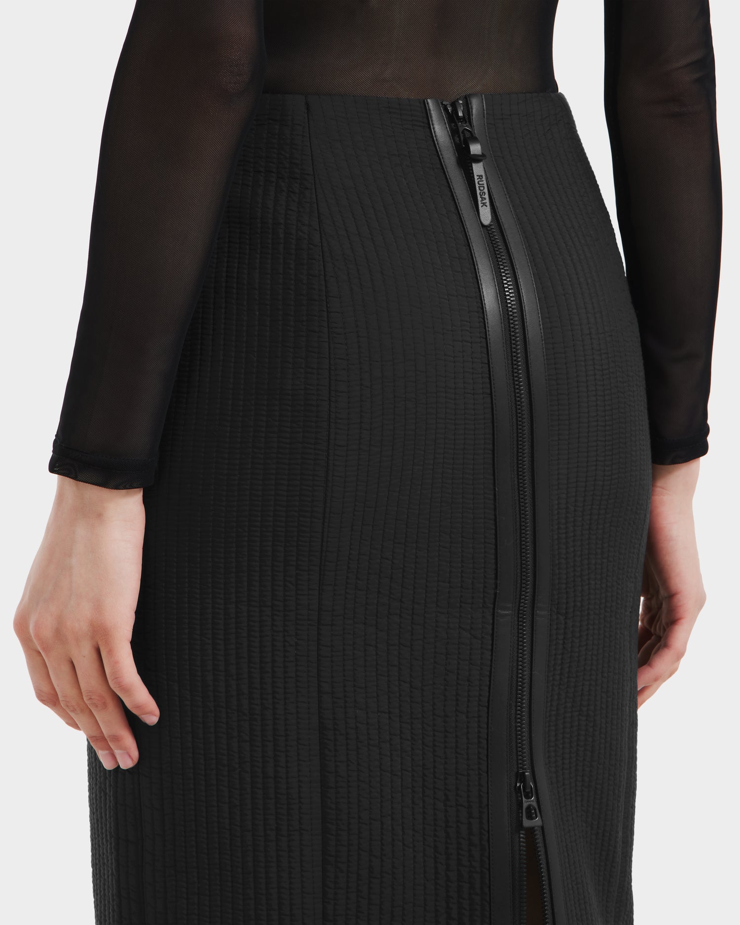 Wearing the Rudsak GLENDORA - 8525563 Black vegan leather fitted skirt with a visible double back zipper, paired with a sheer black long-sleeve top; the image highlights the waist and hips from behind.